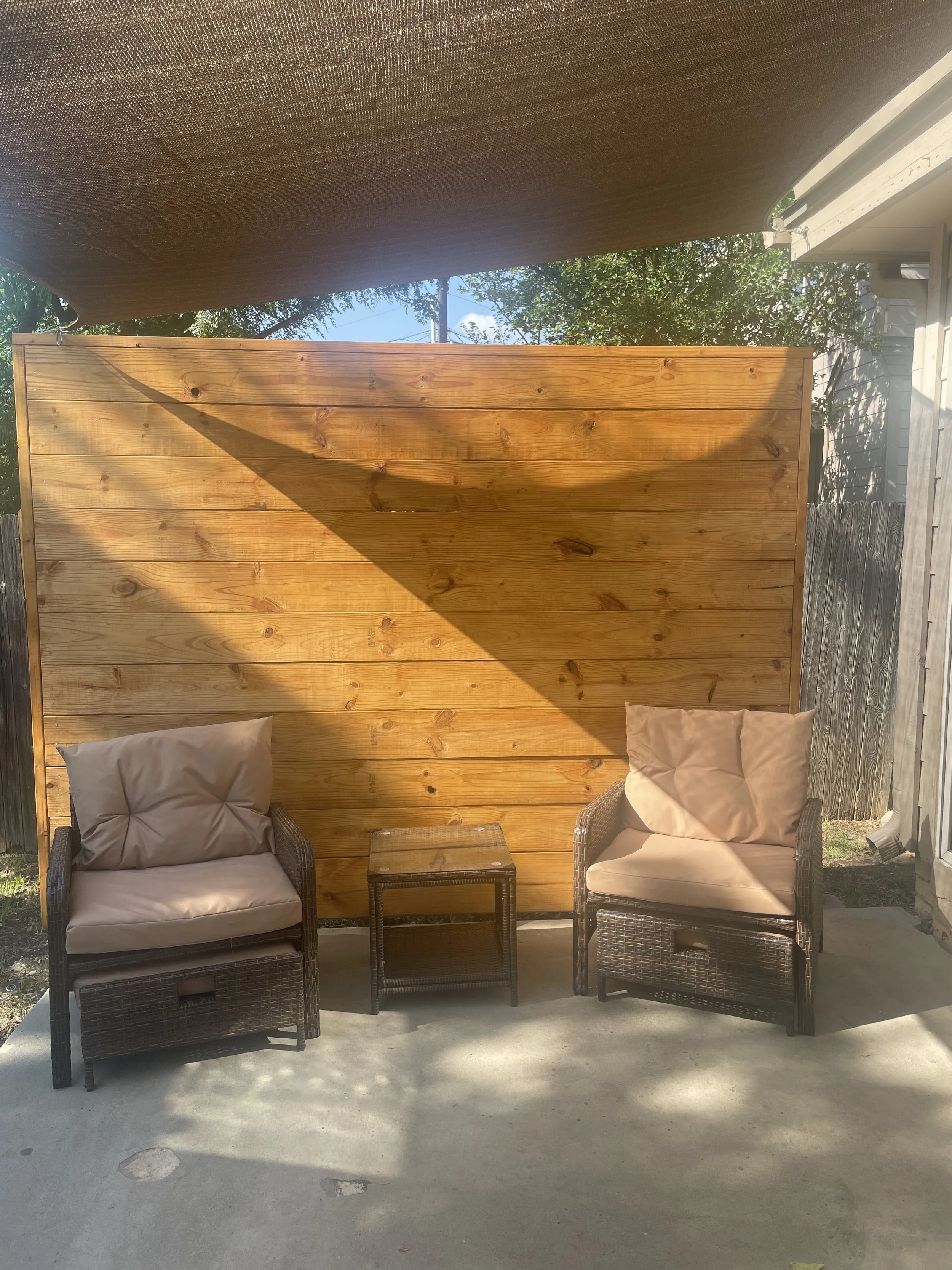 Two outdoor wicker chairs with cushions and a small matching side table set against a wooden privacy wall, with sunlight and shadows cast on the scene.