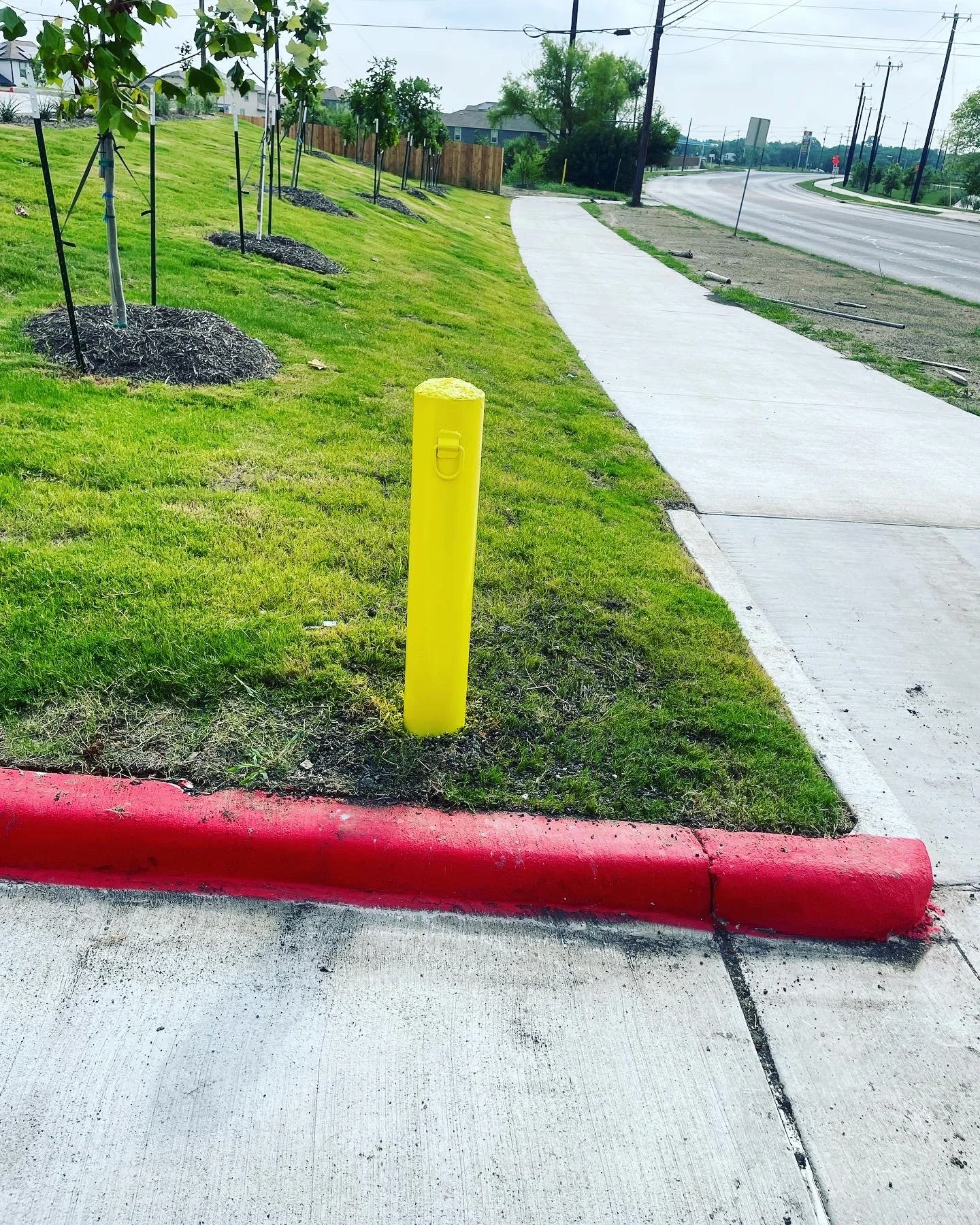 A bright yellow safety bollard that we installed next to a red curb on a sidewalk with grass and trees nearby, with an empty street and utility poles in the background.