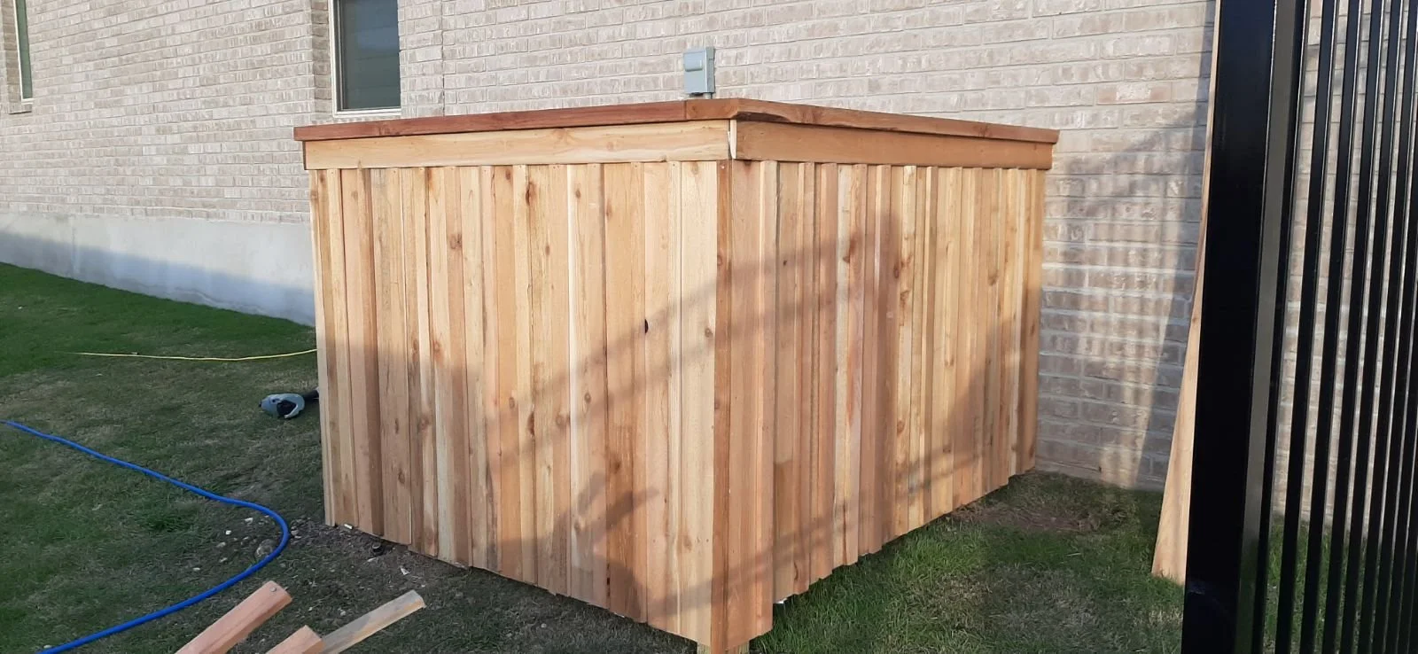 A wooden outdoor trashcan enclosure that we built next to a brick house with a window, on a grassy area with some tools and cords on the ground.