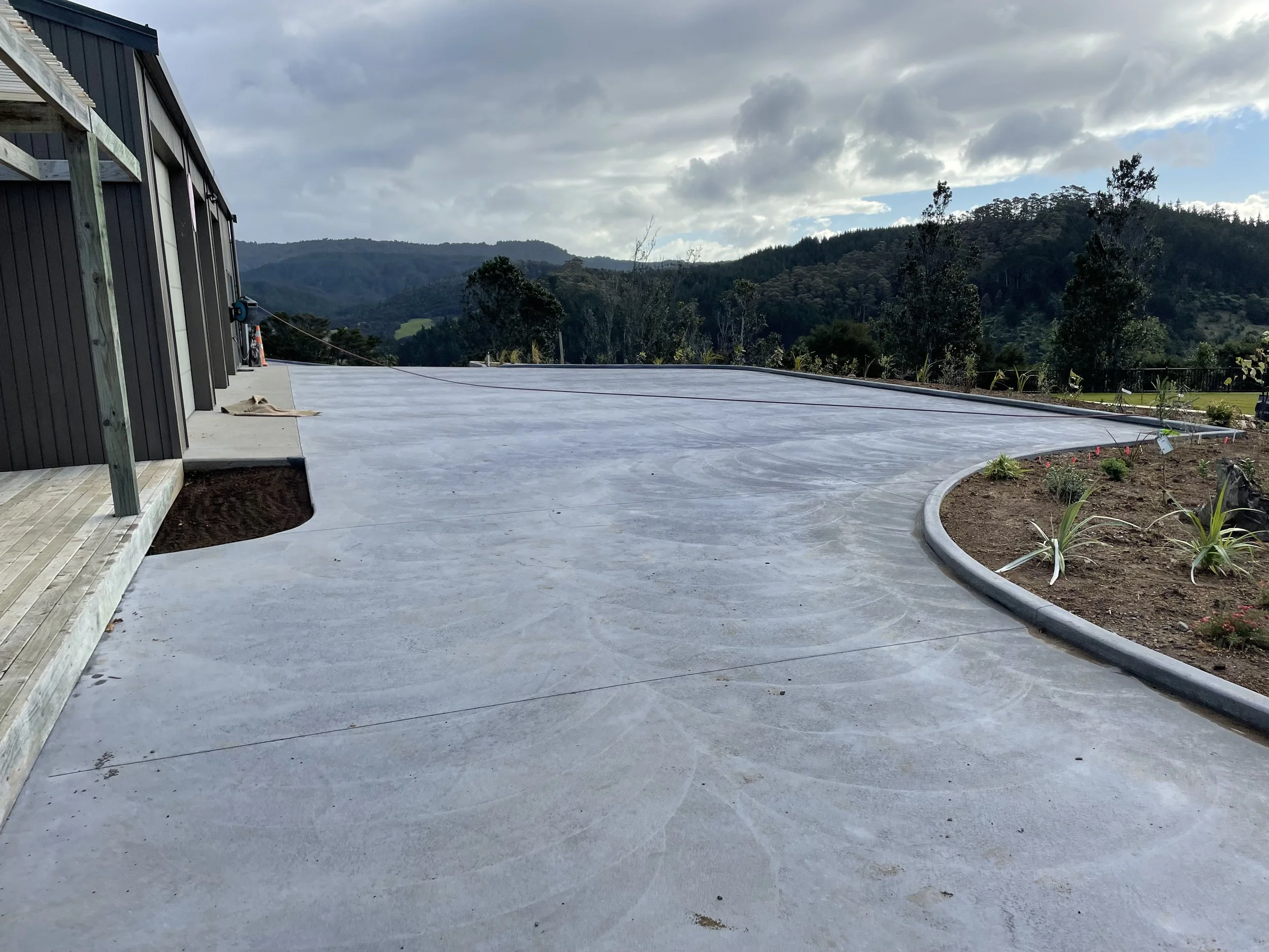Freshly poured concrete driveway or patio area on a sloped terrain, with a curved edge next to a garden bed, and a mountainside with trees and cloudy sky in the background.
