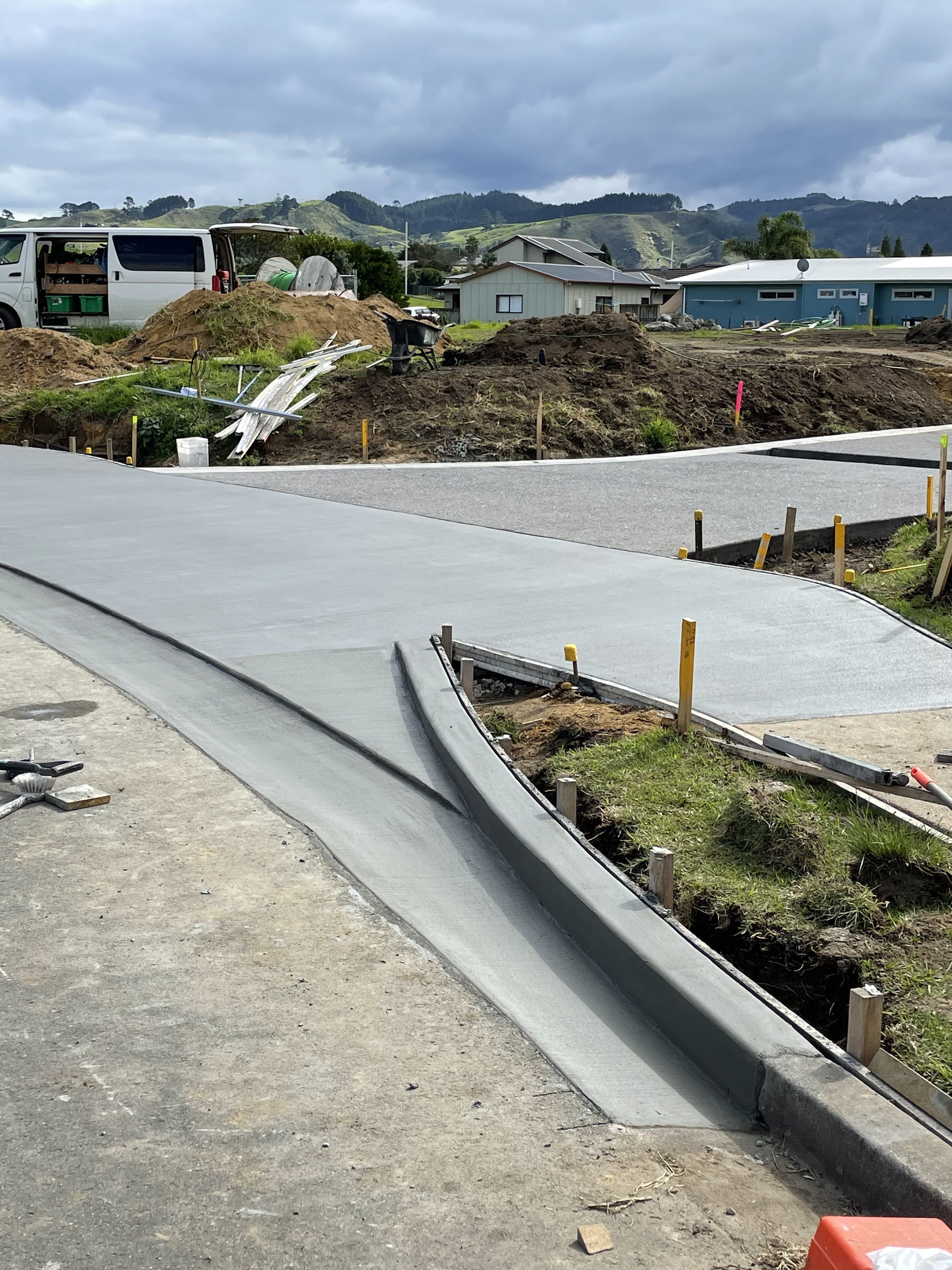 Construction site with freshly poured sidewalks, construction tools, and materials, with houses and hills in the background under cloudy skies.