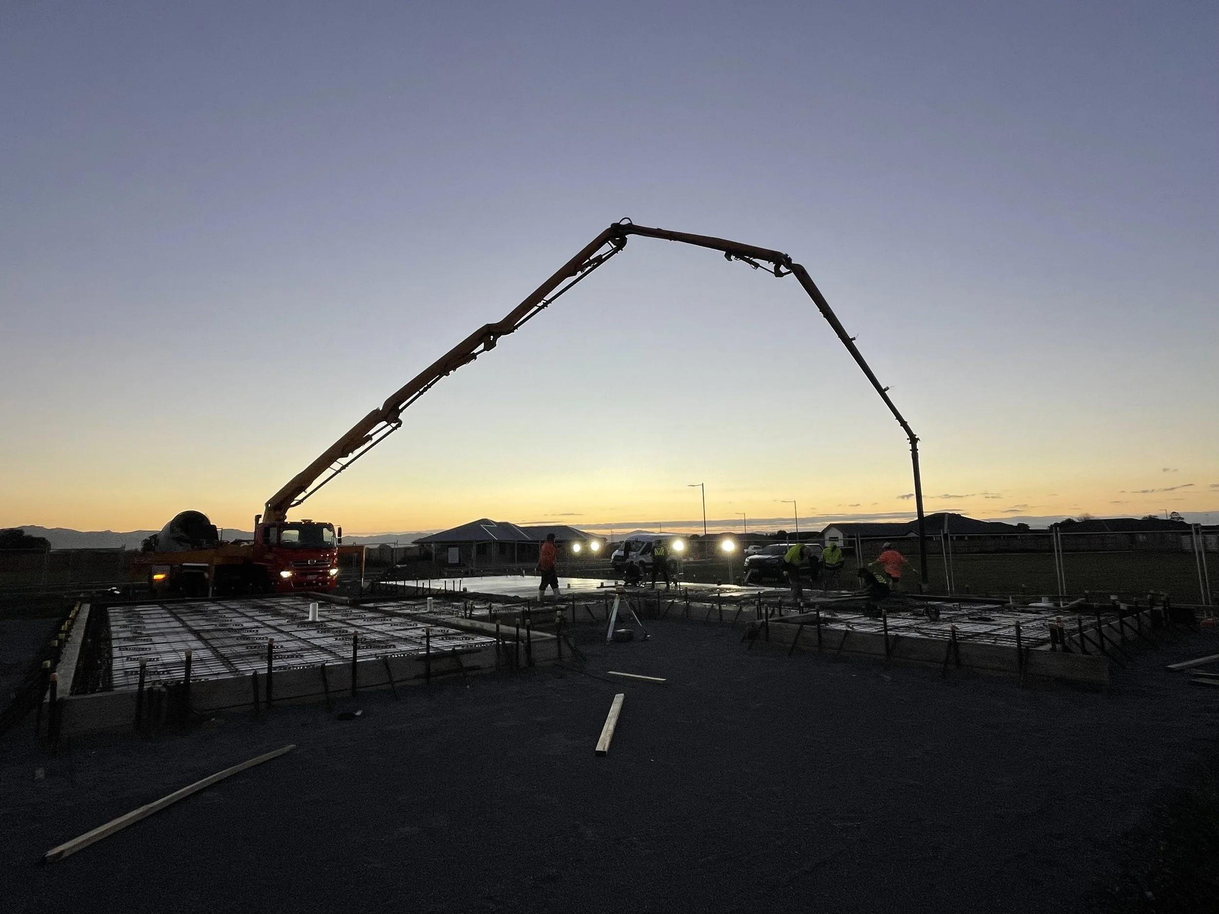 Construction site during dusk with workers, concrete framework, and a concrete pump truck pouring concrete.