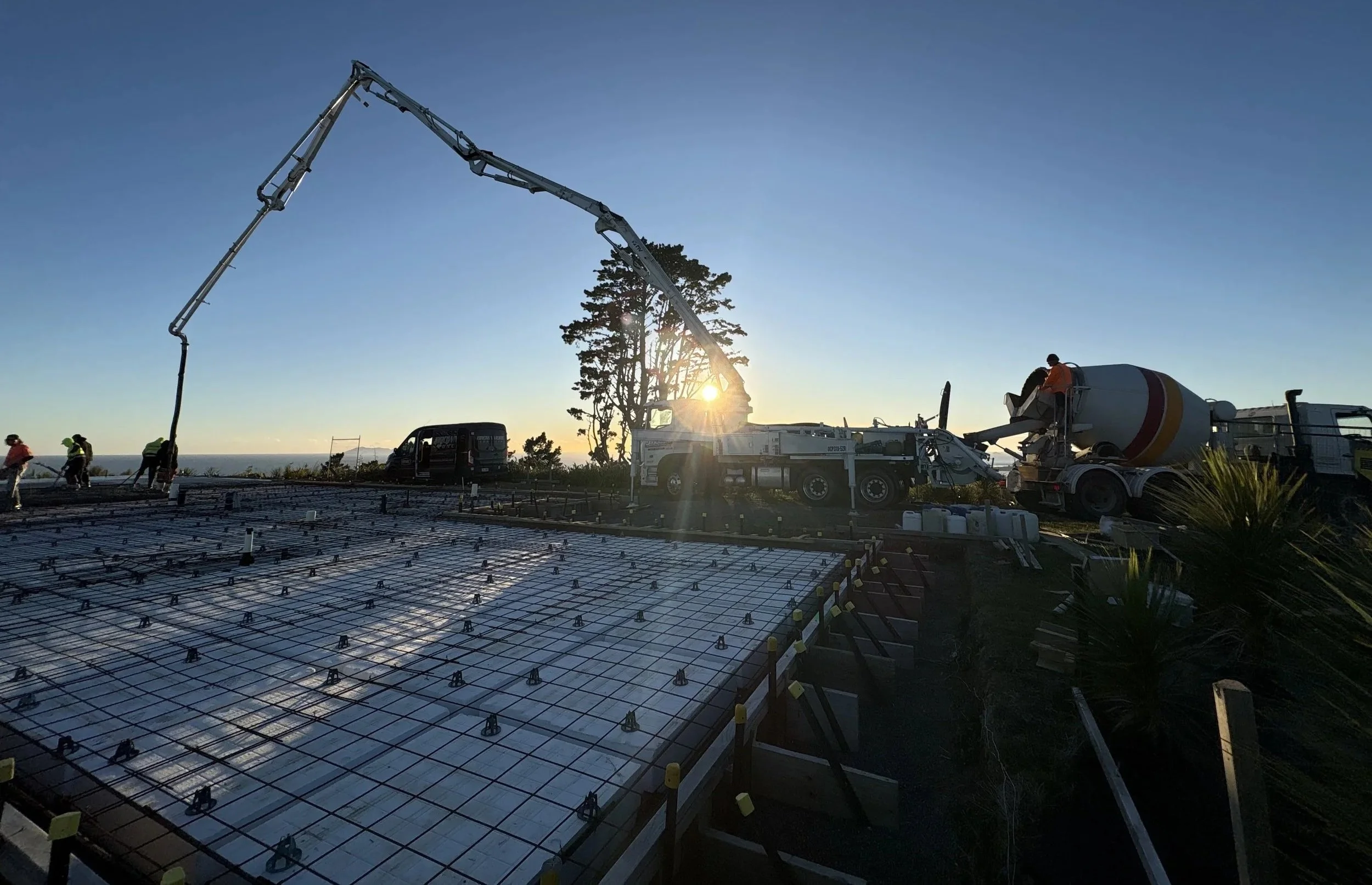Construction workers pouring concrete onto a rebar foundation with a large concrete pump truck at sunset.