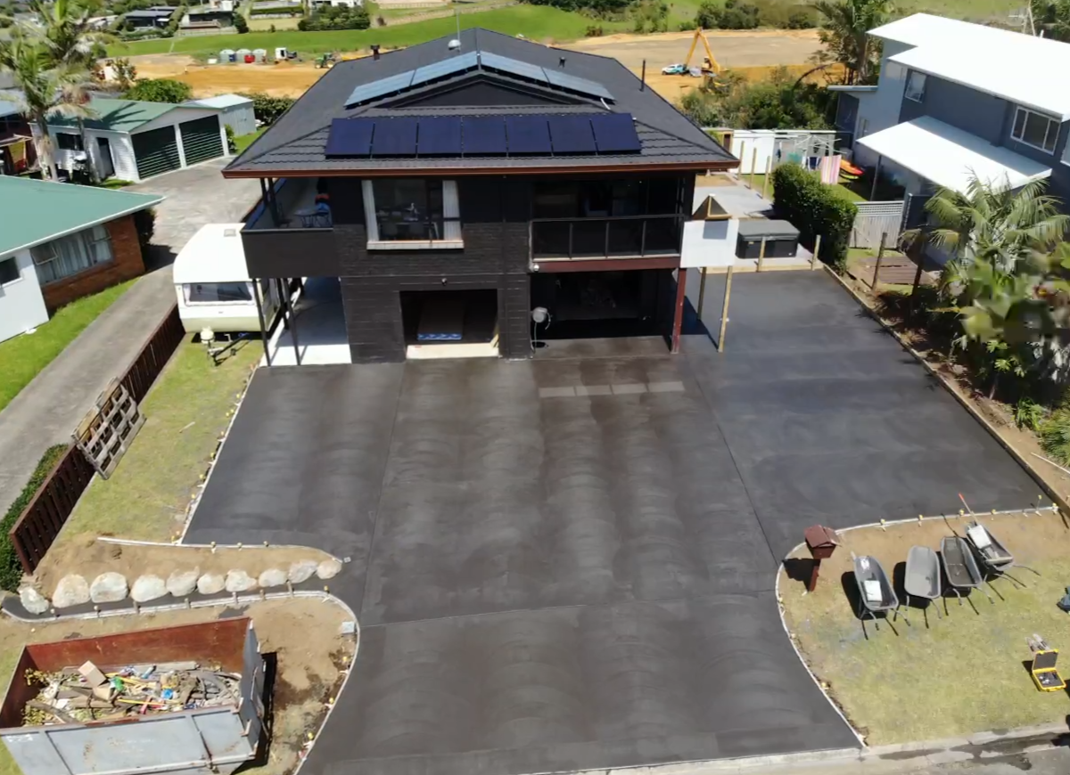 A large house with a newly paved driveway, solar panels on the roof, and a surrounding yard with a trash bin, wheelbarrows, and construction equipment.