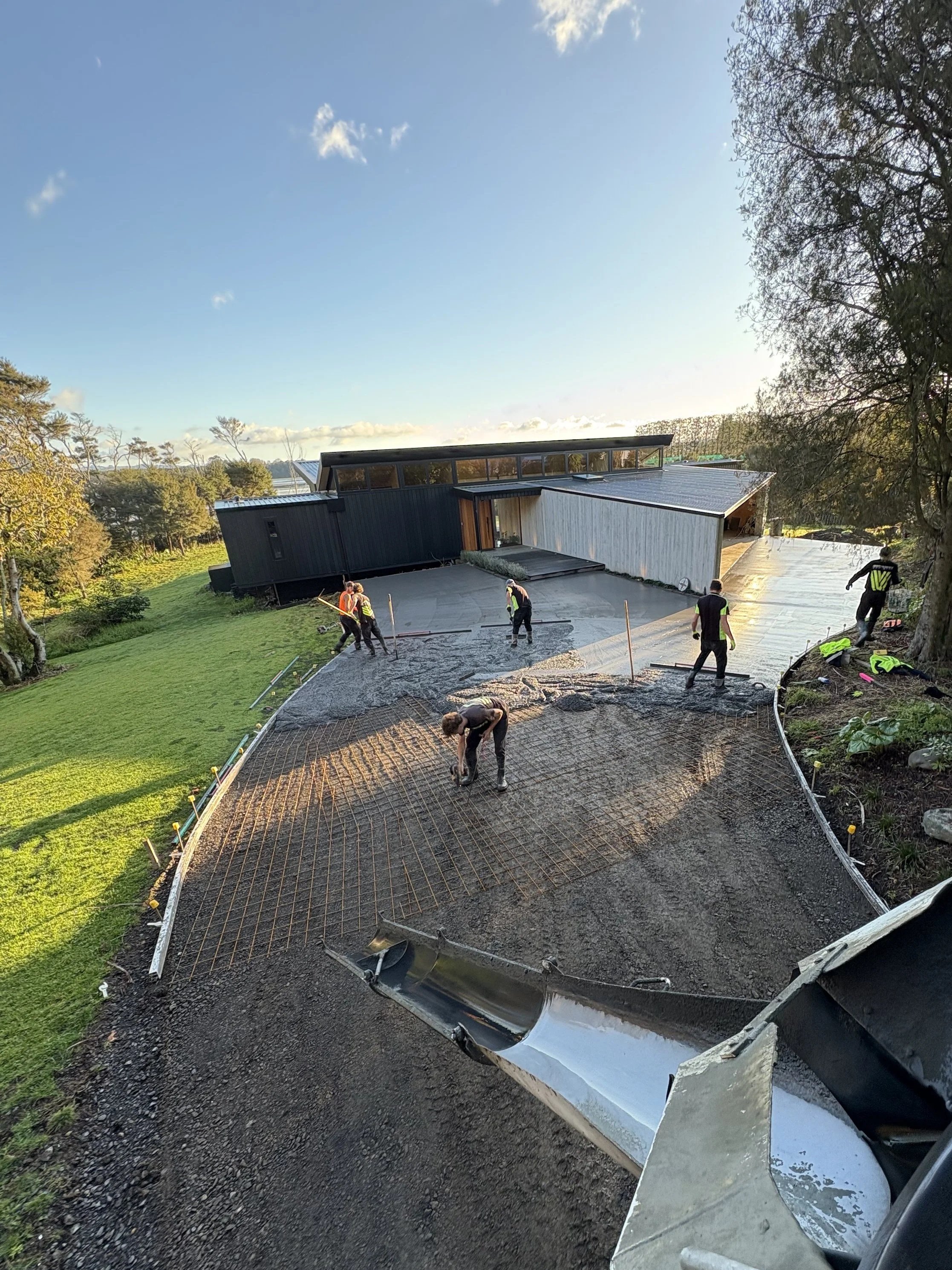 Construction workers pouring and leveling concrete in the front yard of a modern house, surrounded by greenery with a clear sky in the background.