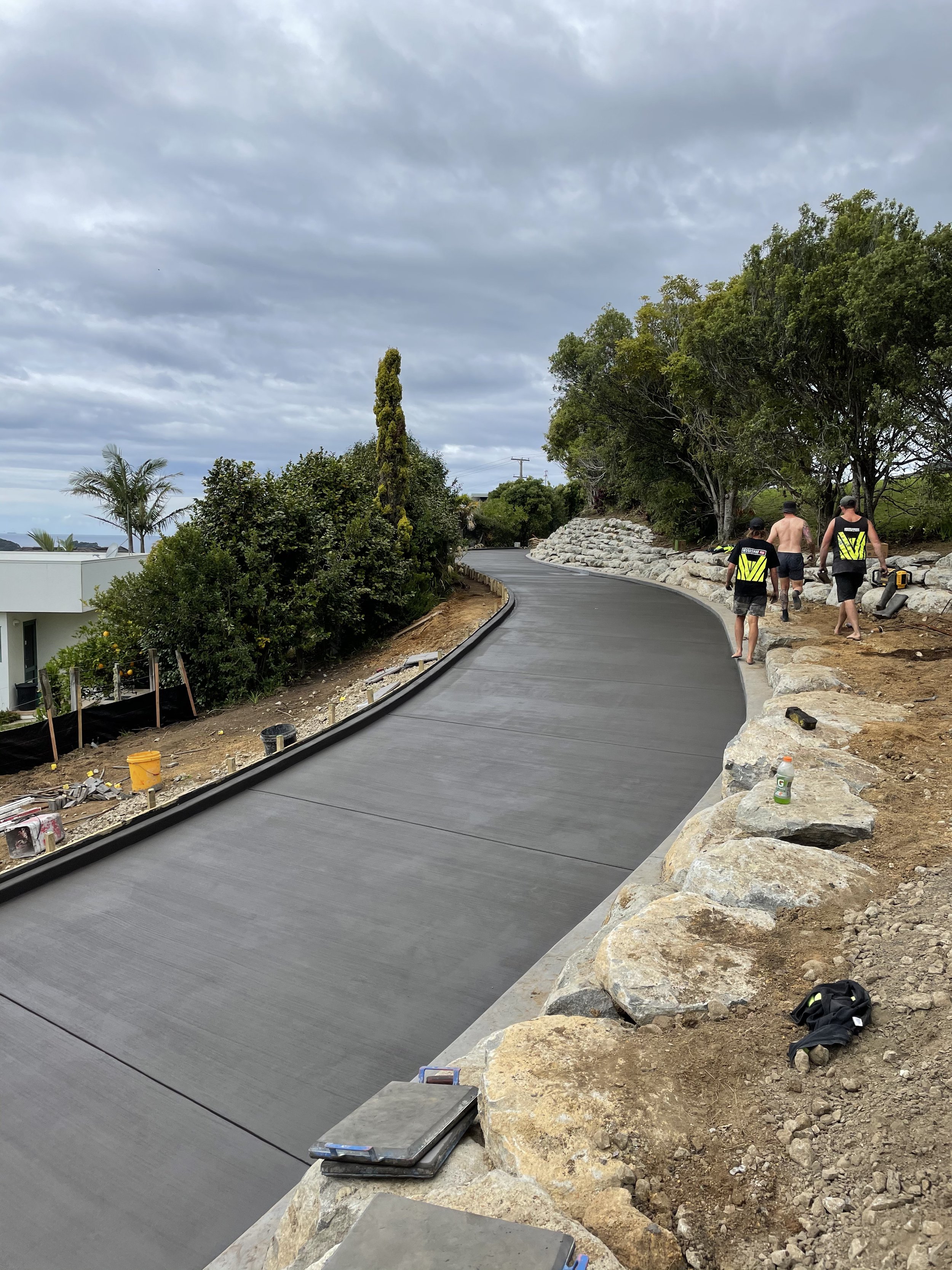 Workers installing a new concrete driveway or pathway with rocks and trees on the side, under cloudy skies.