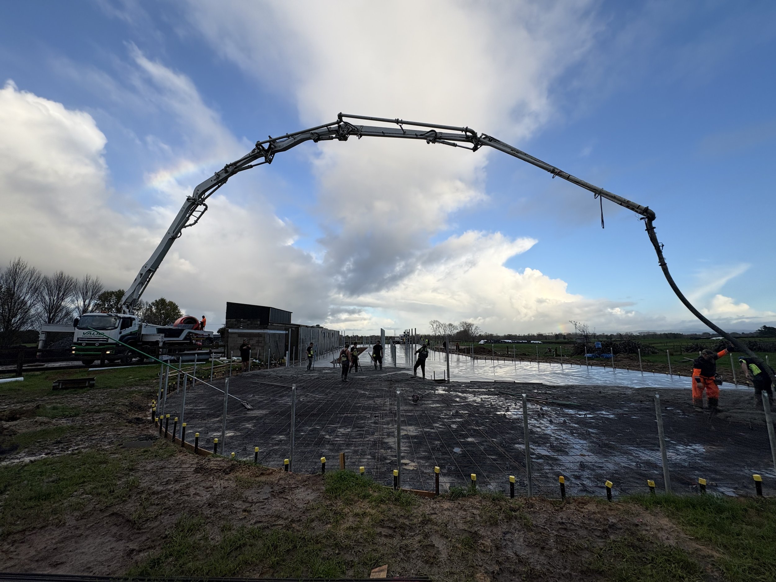 Construction workers pouring concrete for a building foundation with a concrete pump truck extending its arm over the site. The sky is partly cloudy, and the site is fenced off.