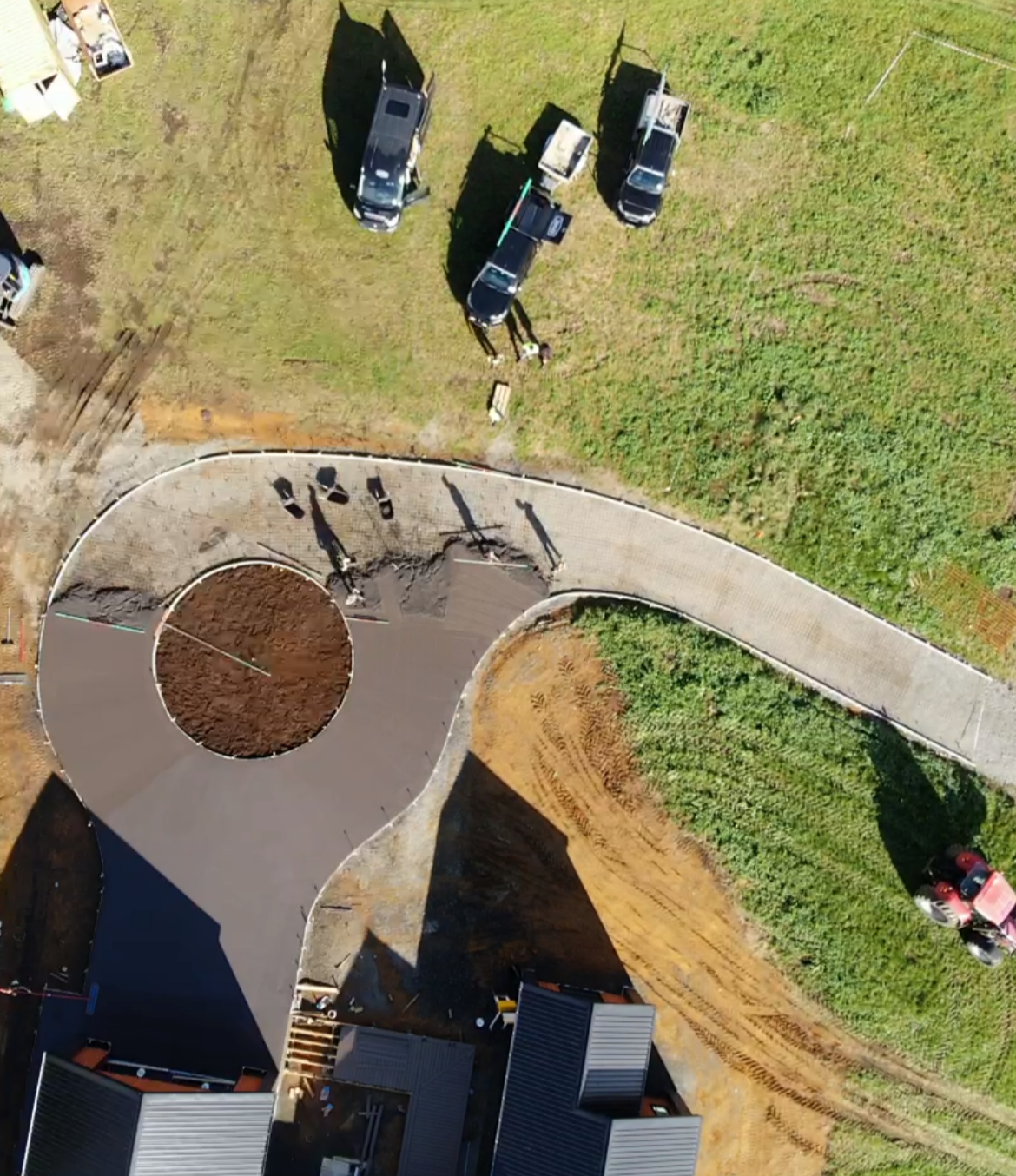 An aerial view showing a construction site with a curved driveway, several parked cars, and construction materials. The site is surrounded by grassy areas and some buildings with metal roofs.