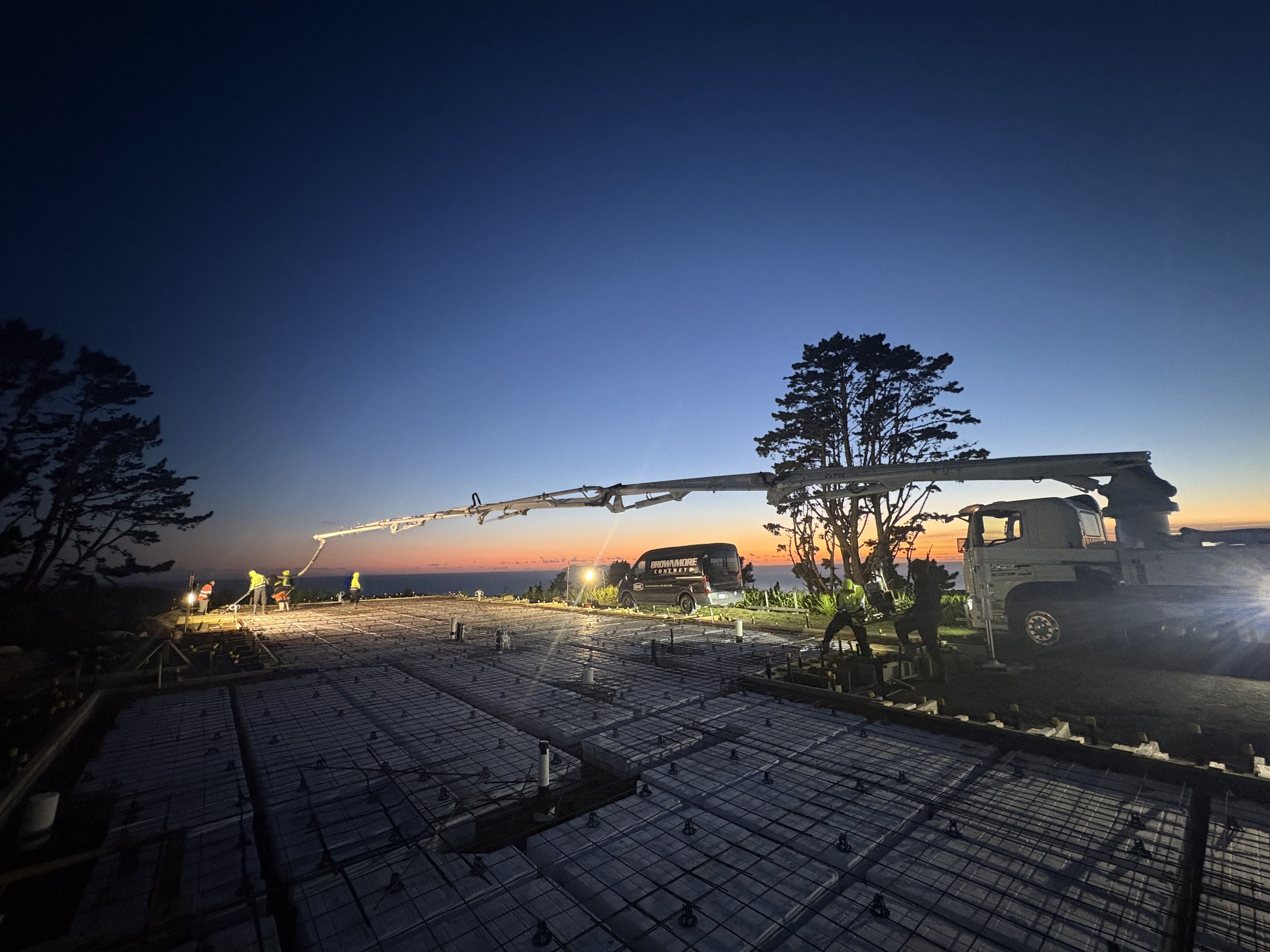 Construction workers working on a building foundation at dusk with a concrete pump truck and trees in the background.