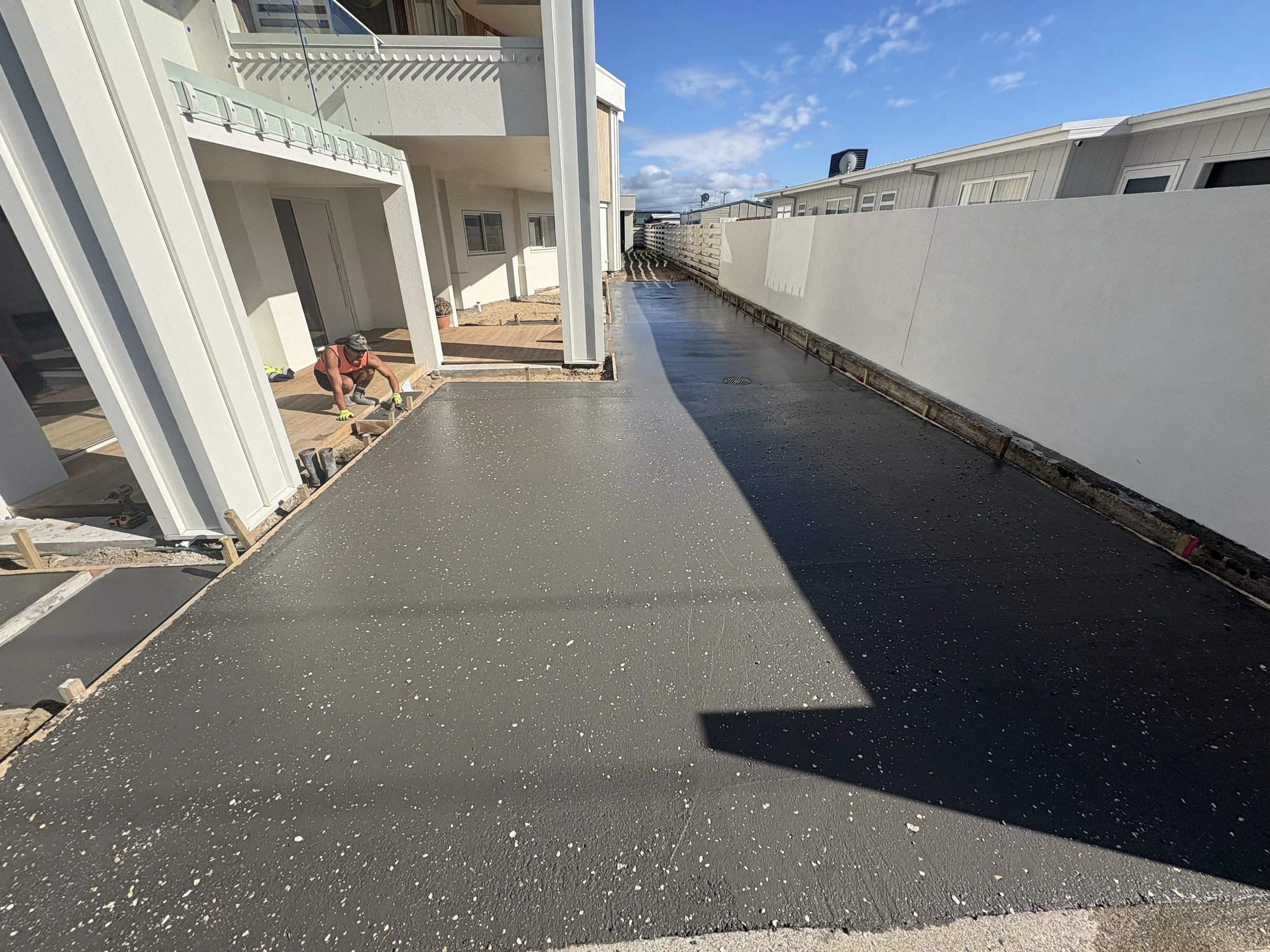 Construction worker applying fresh black asphalt on a patio in a residential area, with the sun casting shadows on the ground and modern white buildings nearby.