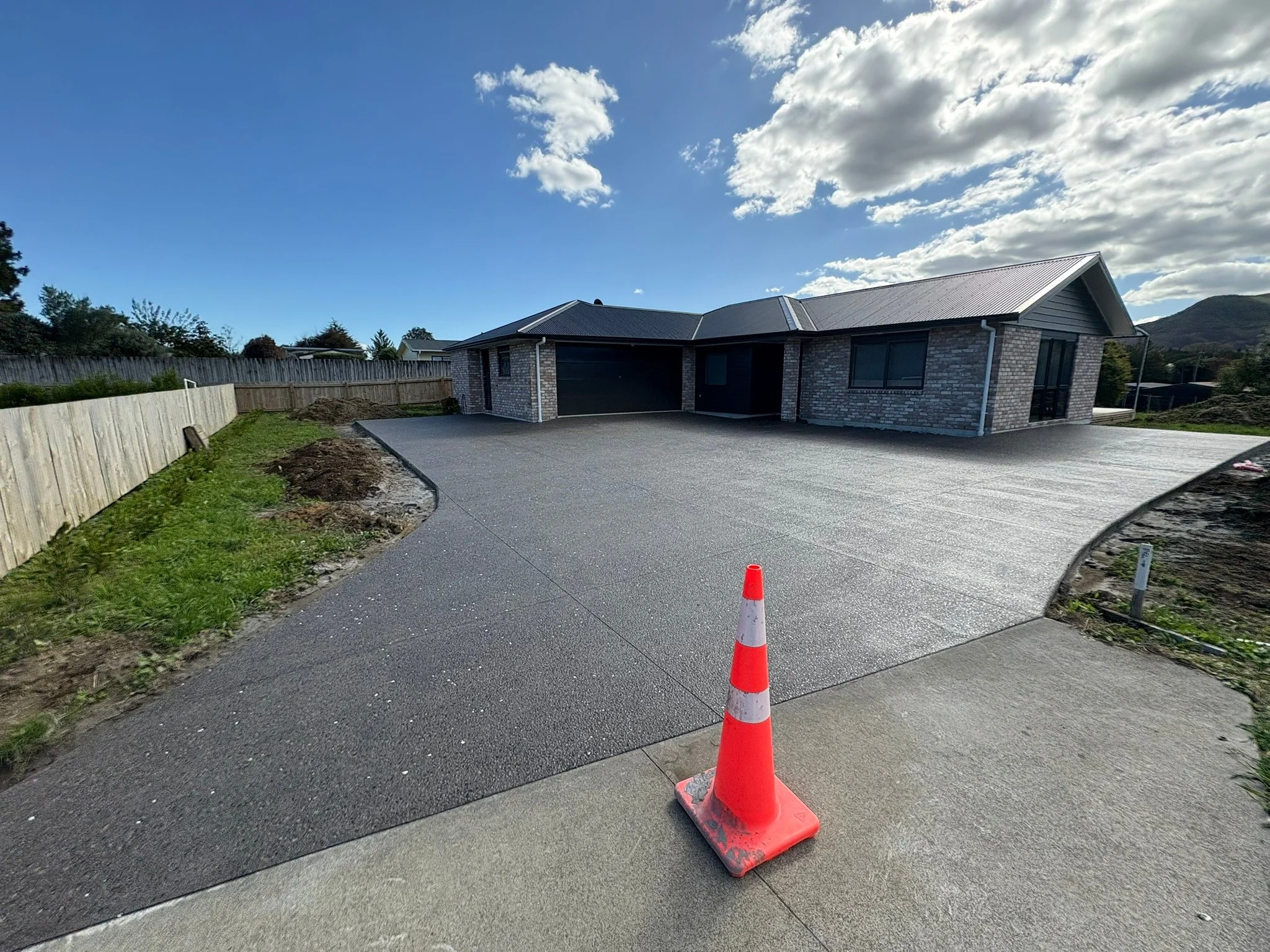 Newly paved driveway in front of a modern house with brick exterior, with a construction cone placed on the sidewalk, and a blue sky with some clouds above.
