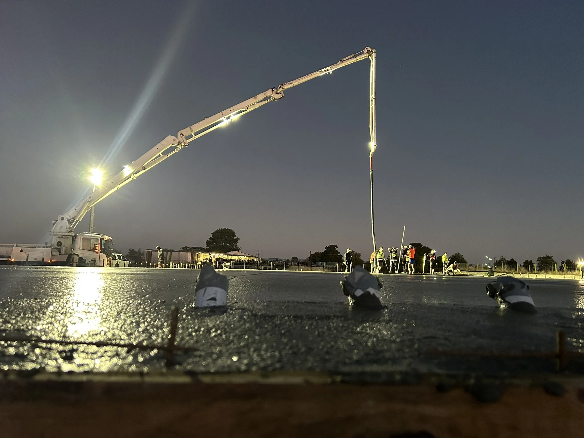 Workers at a construction site pouring asphalt during nighttime with a large crane overhead and multiple workers in safety gear.