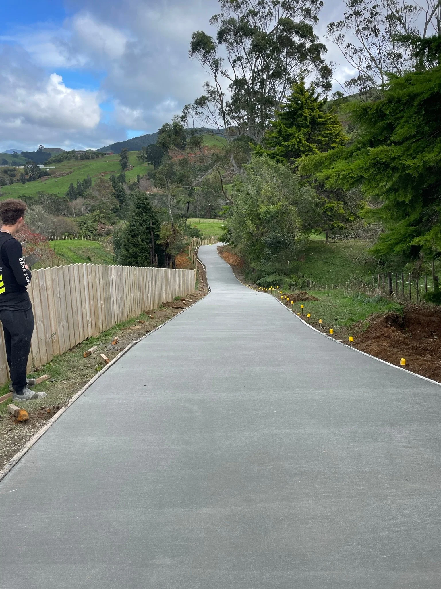 A newly paved concrete walking path winding through a hilly, rural landscape with trees and fields, under a partly cloudy sky. A person stands on the left side, observing the path.