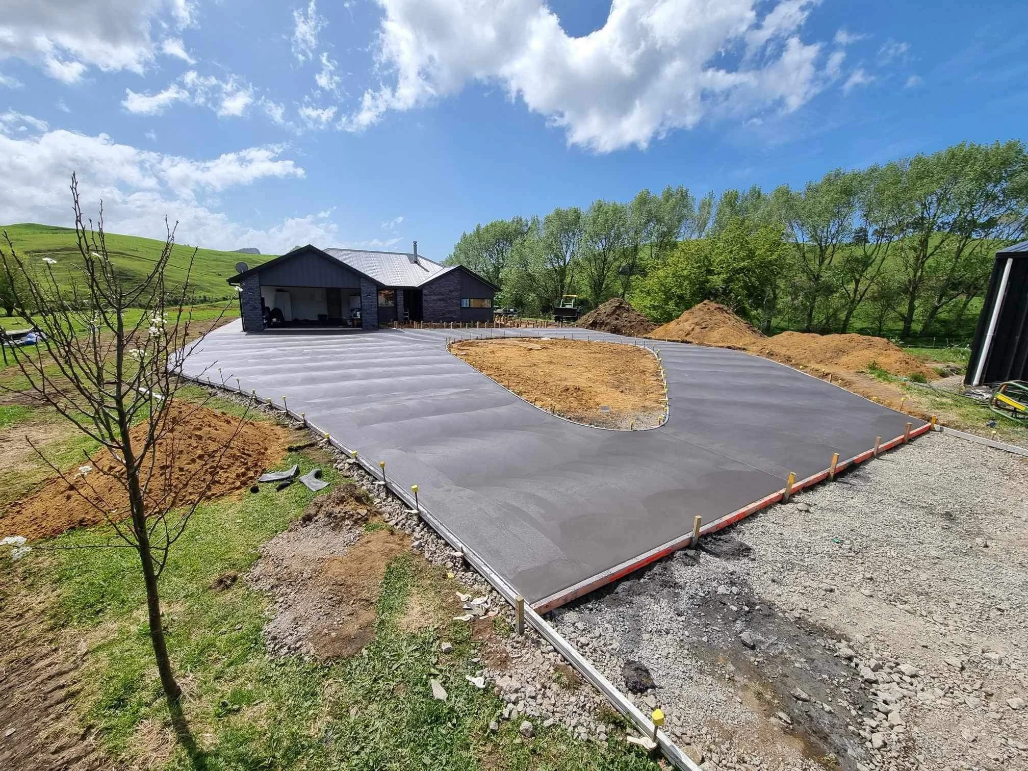 Newly paved concrete driveway in the process of being completed, with form boards and stakes in place, a house in the background, and piles of dirt beside the driveway, surrounded by greenery and trees under a partly cloudy sky.