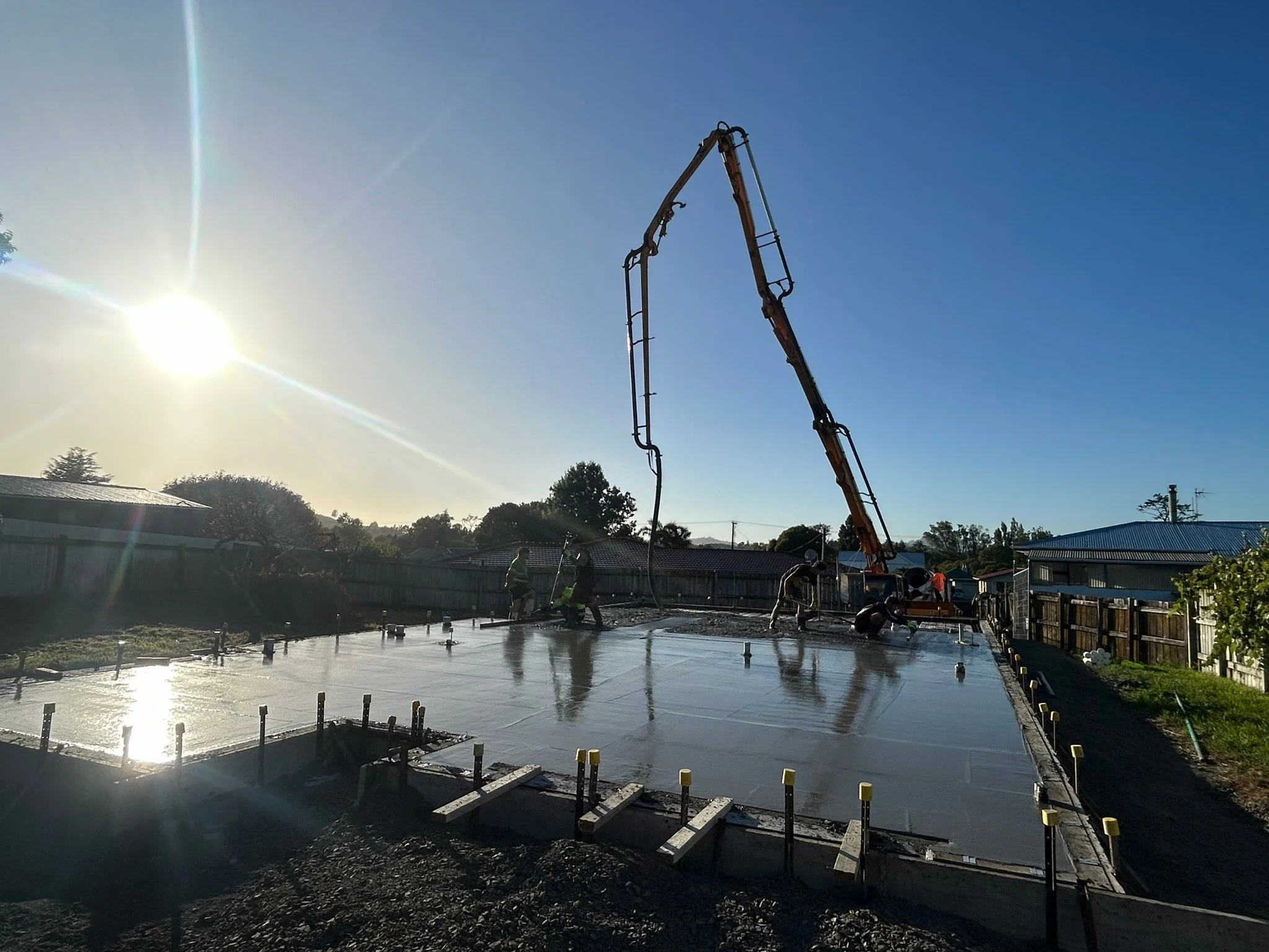 Construction workers pouring concrete on a building foundation with a cement truck and boom pump under a bright sun and clear sky.
