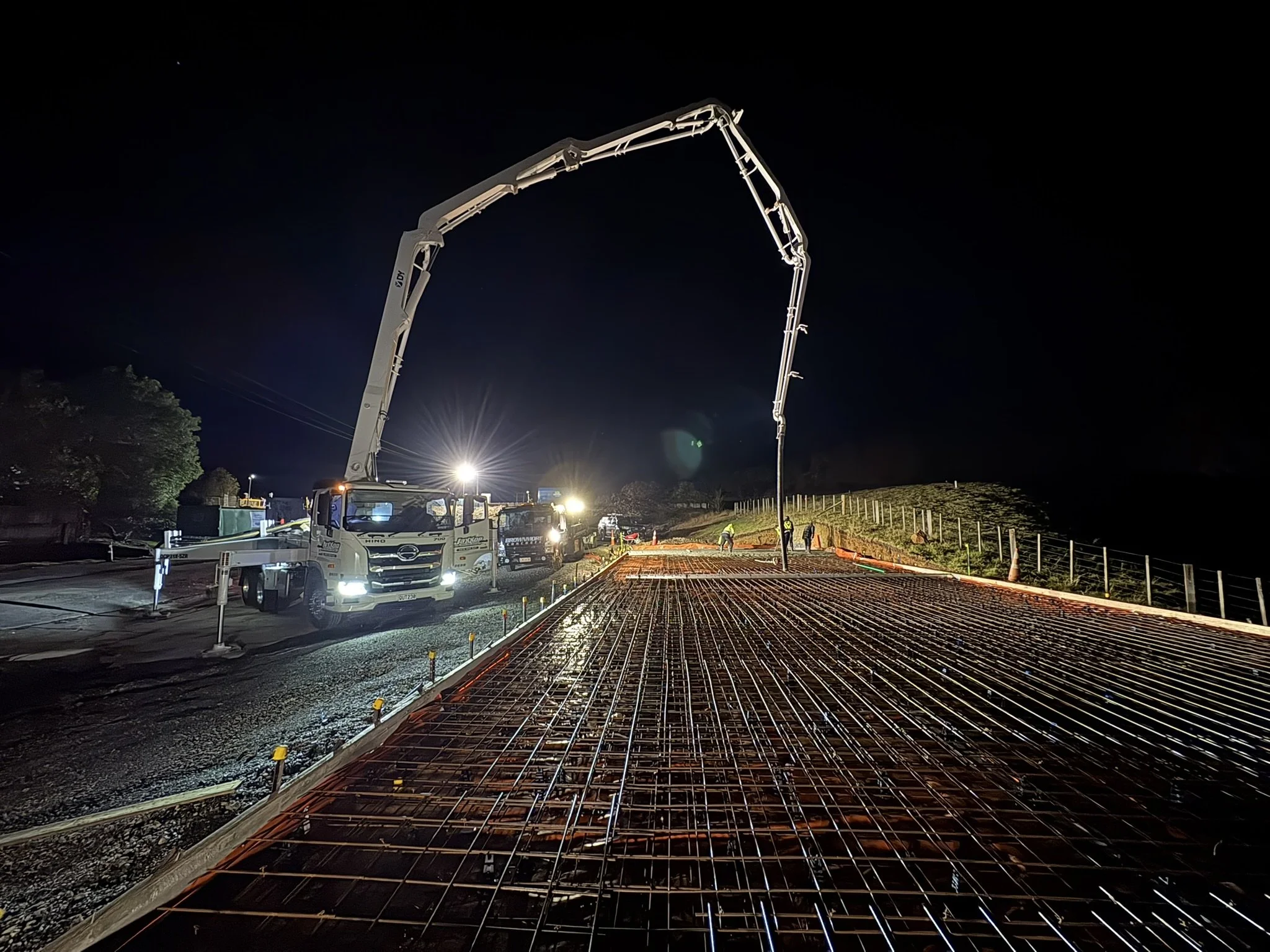 Construction workers pouring concrete on a road at night using a concrete pump truck with an extended boom.