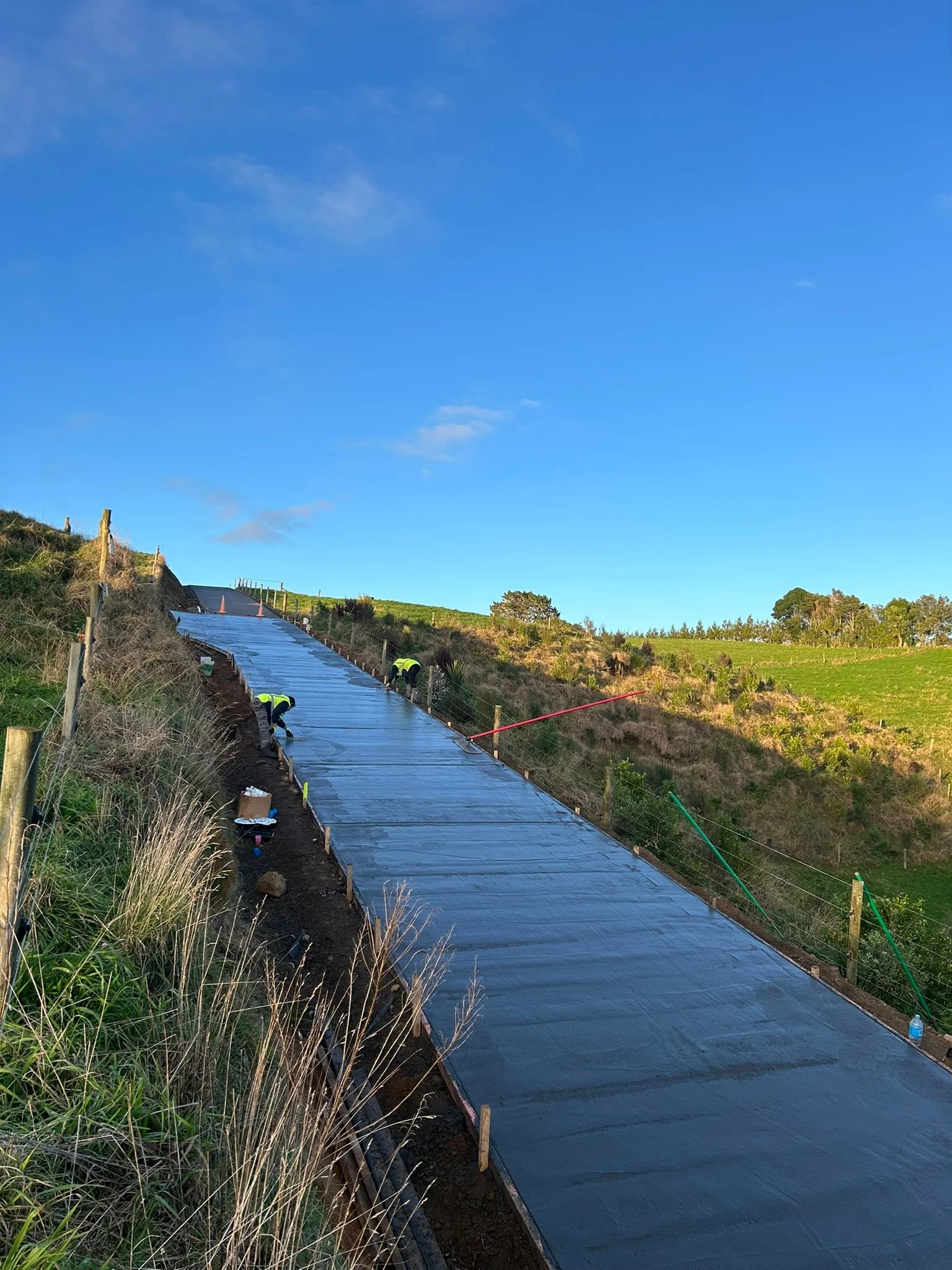 Construction workers laying fresh concrete on a sloped outdoor pathway or road on a hillside under a clear blue sky.