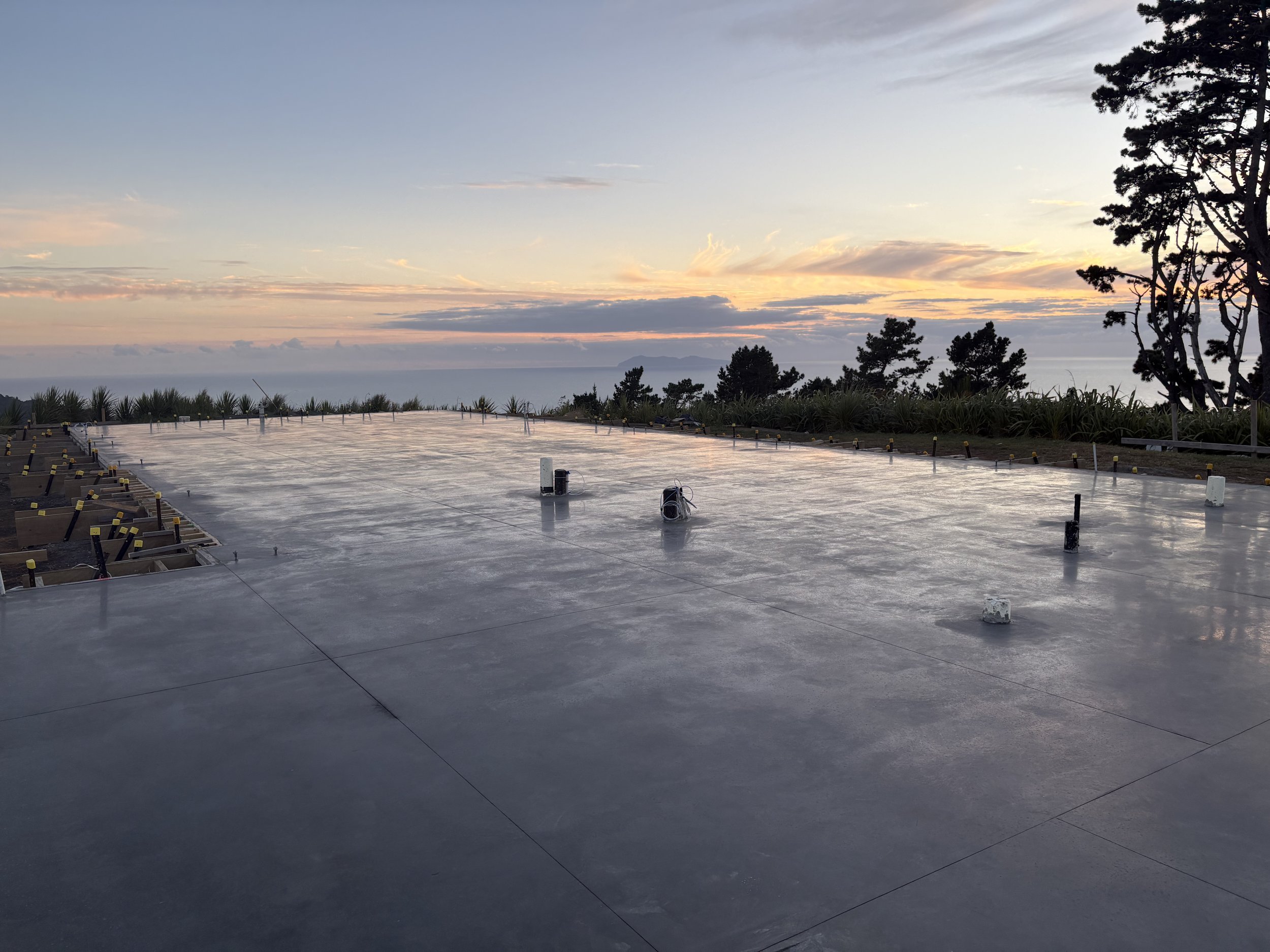 Construction site with a newly poured concrete slab under a sunset sky, surrounded by trees and shrubbery.