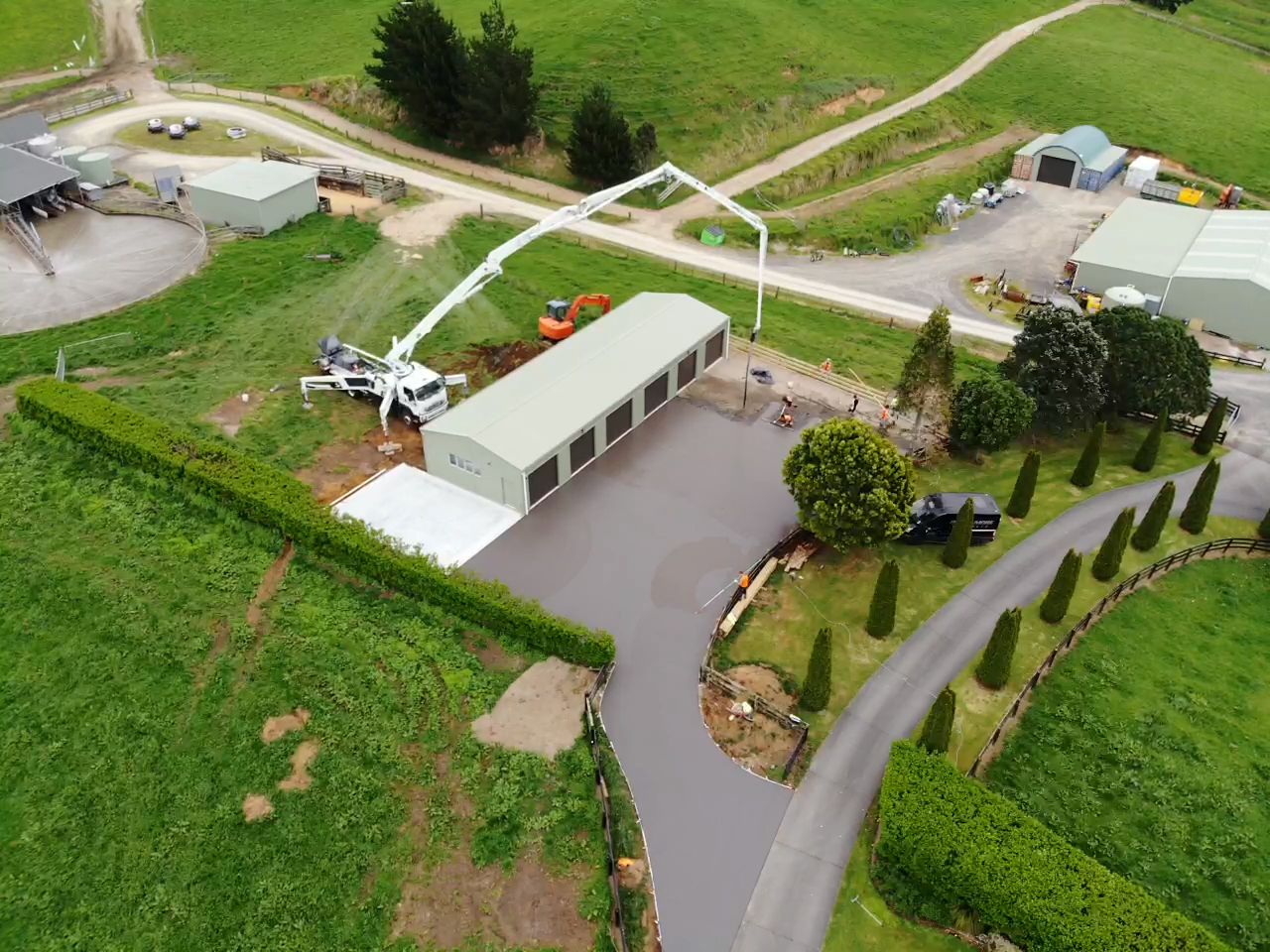 An aerial view of a construction site with a concrete pump truck pouring concrete for a new building, surrounded by green grass, trees, and nearby buildings and roads.