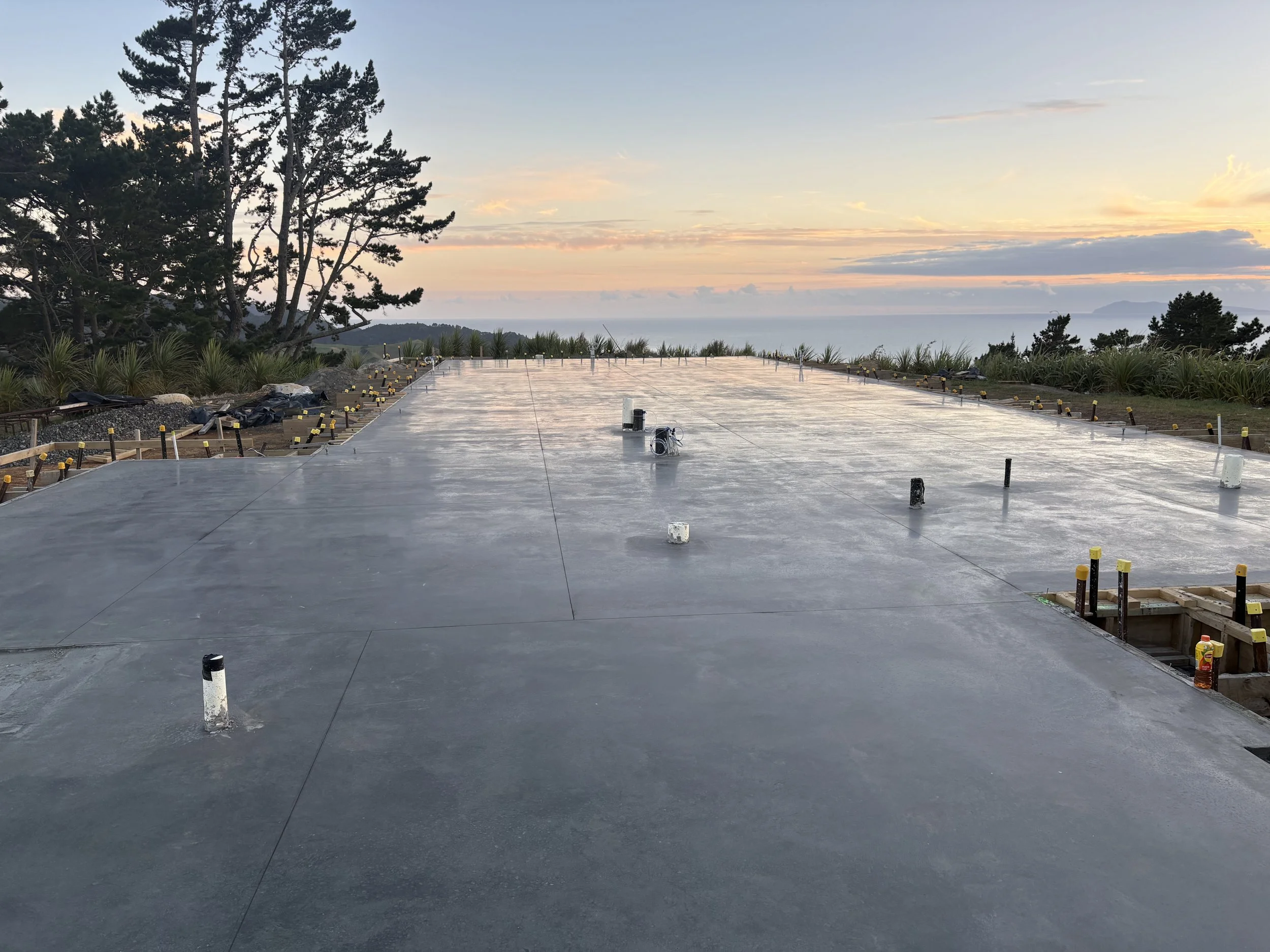 Construction site with a newly poured concrete slab in an outdoor area, with trees and a sunset sky in the background.