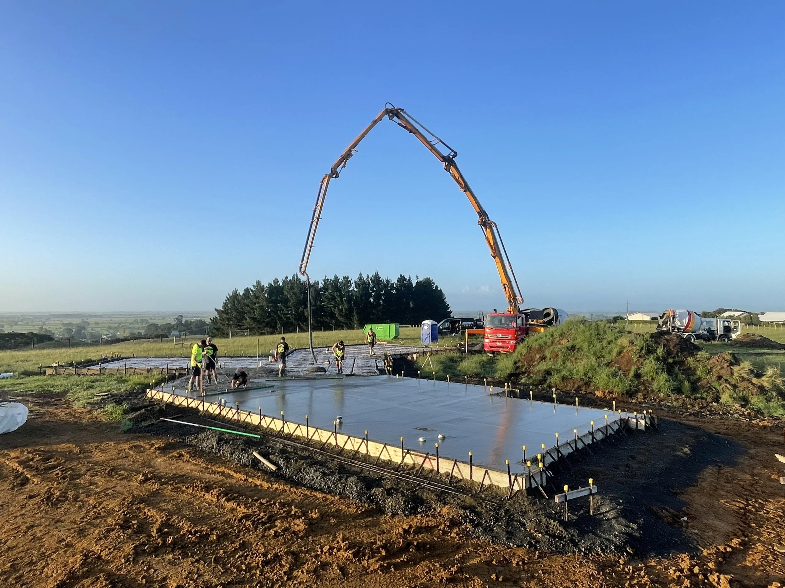 Construction workers with high-visibility vests working on pouring a concrete foundation on a construction site, with a concrete pump truck in operation during daylight hours.