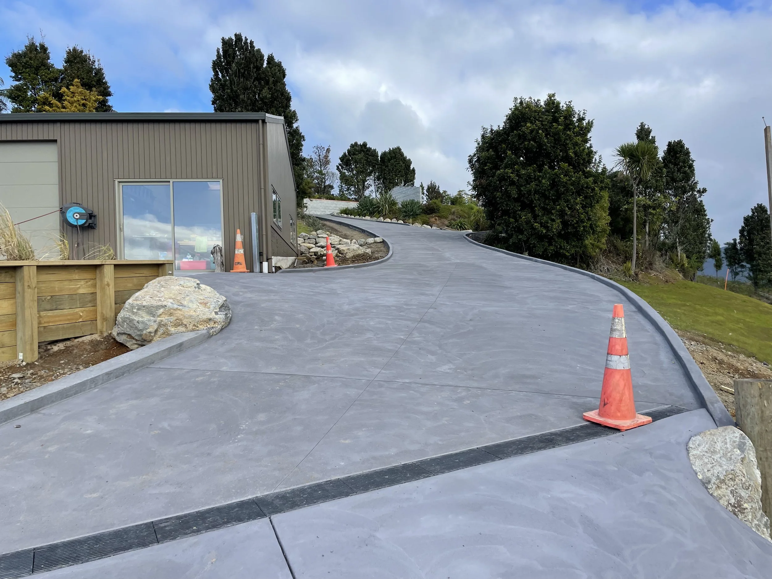 Newly paved, curved concrete driveway leading to a building with gray siding, surrounded by trees and a grassy hill, with construction cones placed on the side.