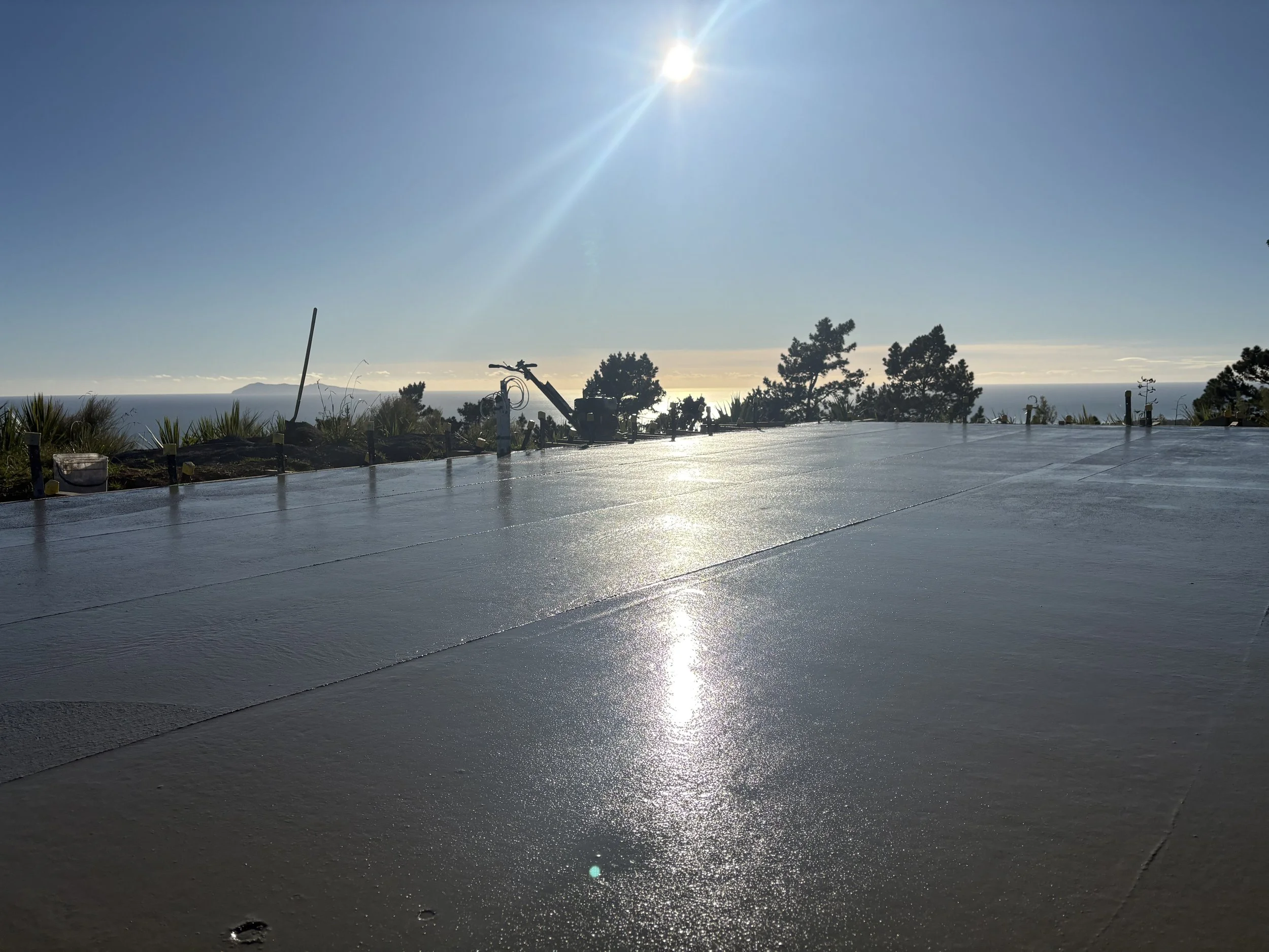 Sunny outdoor scene with a flat rooftop surface, trees in the background, and the ocean visible beyond with the sun shining brightly.