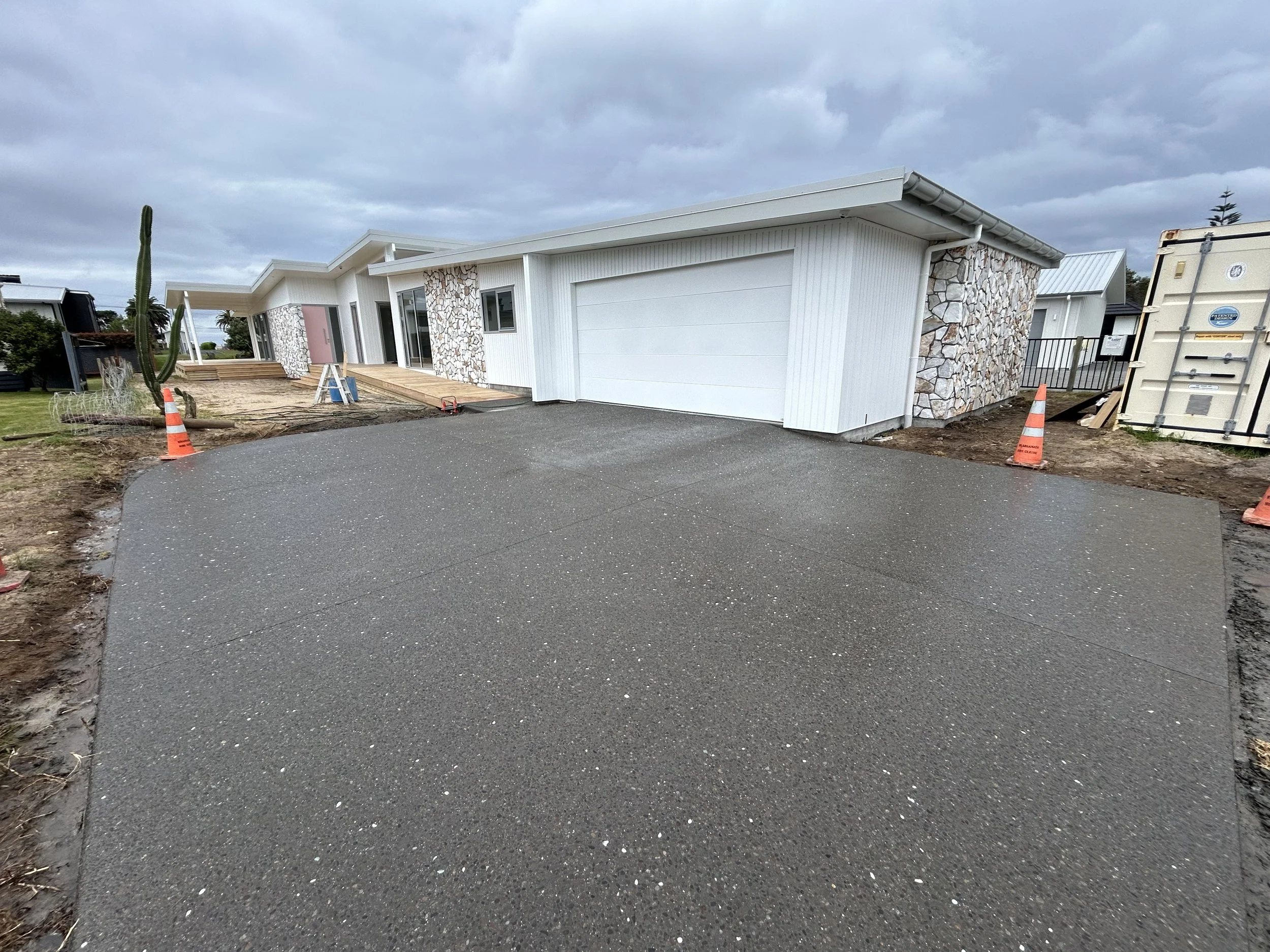 Front view of a modern house under construction with a smooth, freshly poured concrete driveway, construction cones, and a cloudy sky.
