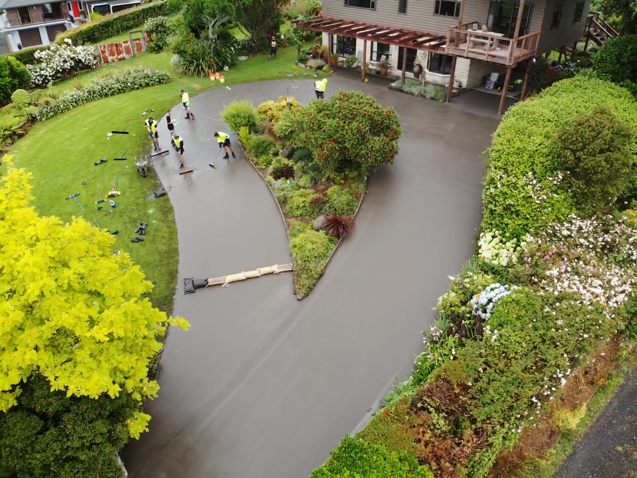 Aerial view of a residential house with a newly paved driveway, small garden, and workers installing the driveway surface.