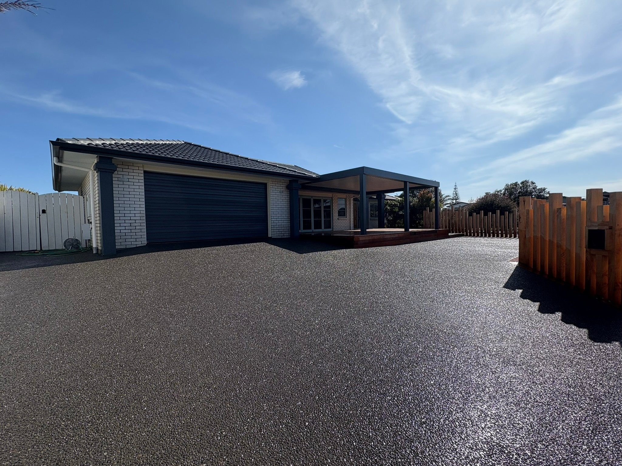 A modern house with a garage, a covered deck area, and a wooden fence on an asphalt driveway under a blue sky.