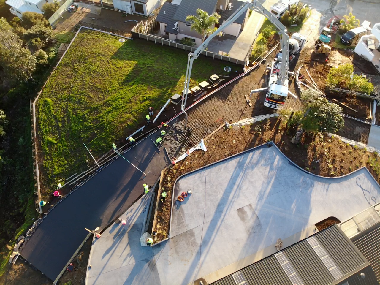 Workers pour concrete onto a curved section of a rooftop construction project, with a concrete mixer truck and a concrete pump in use.