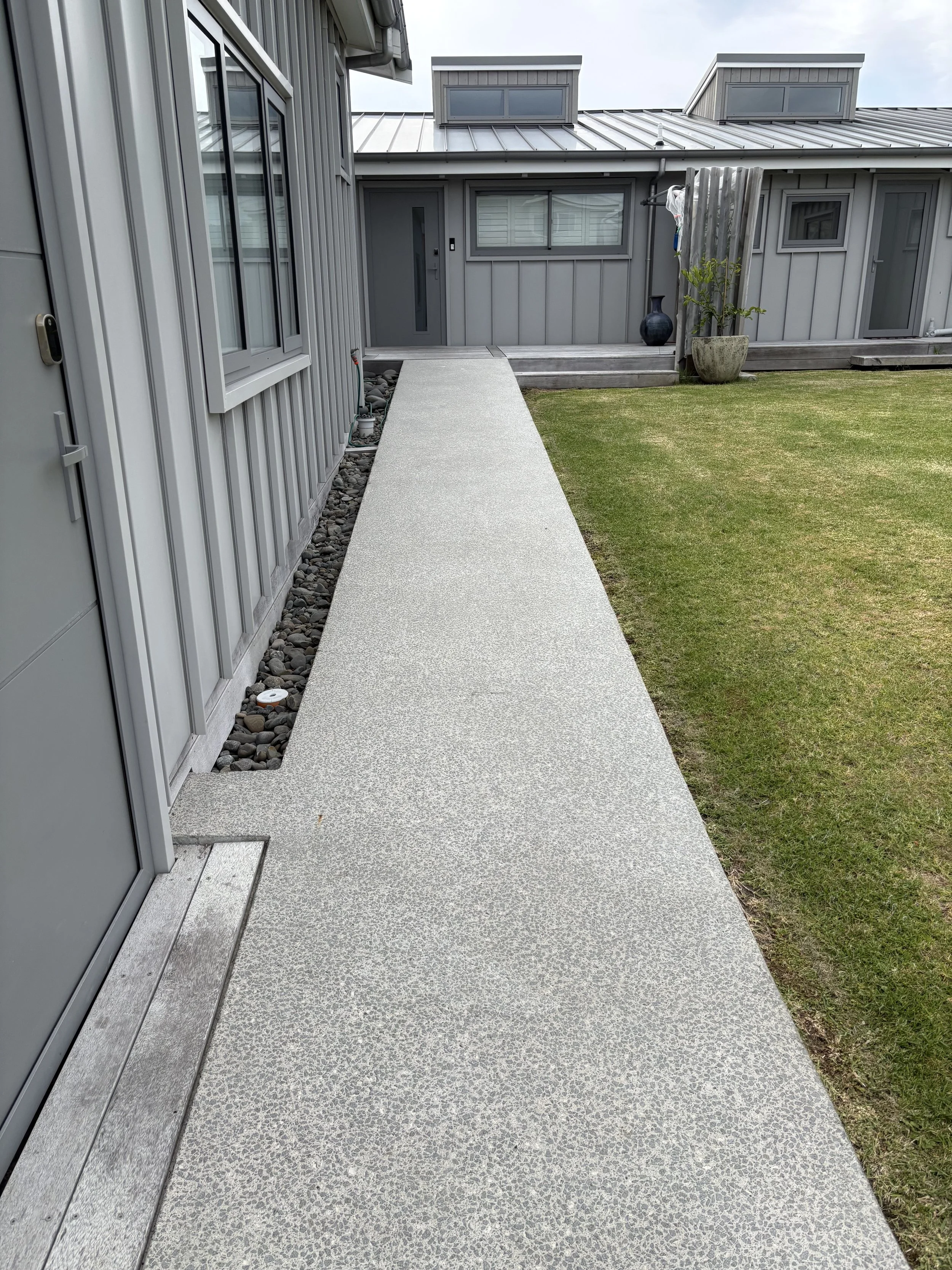 A concrete walkway running alongside a gray house with a window and door, leading to a backyard with a grassy lawn and a gray building in the background.