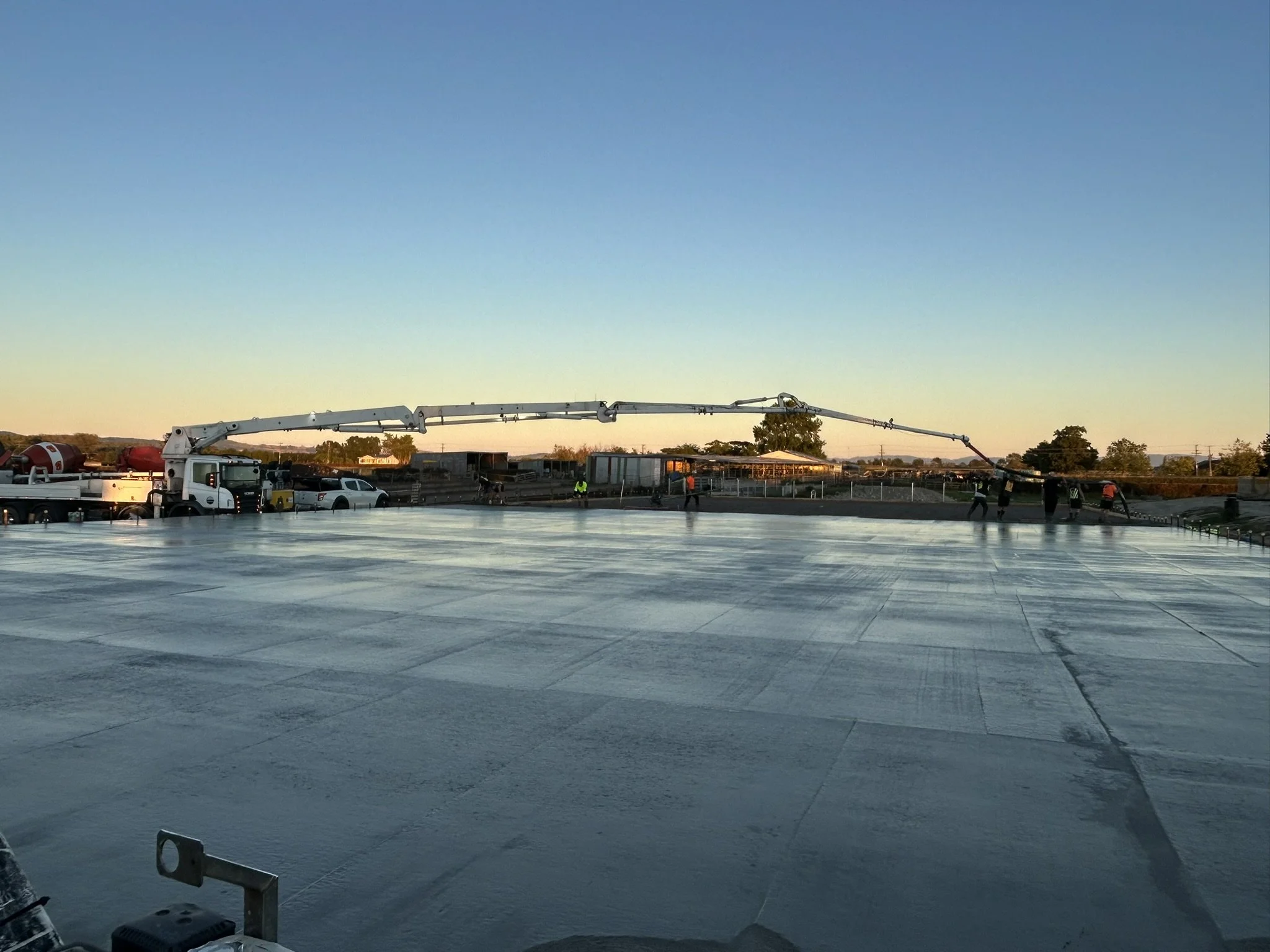 Construction workers pouring concrete on a large flat surface using a concrete pump truck during sunset.