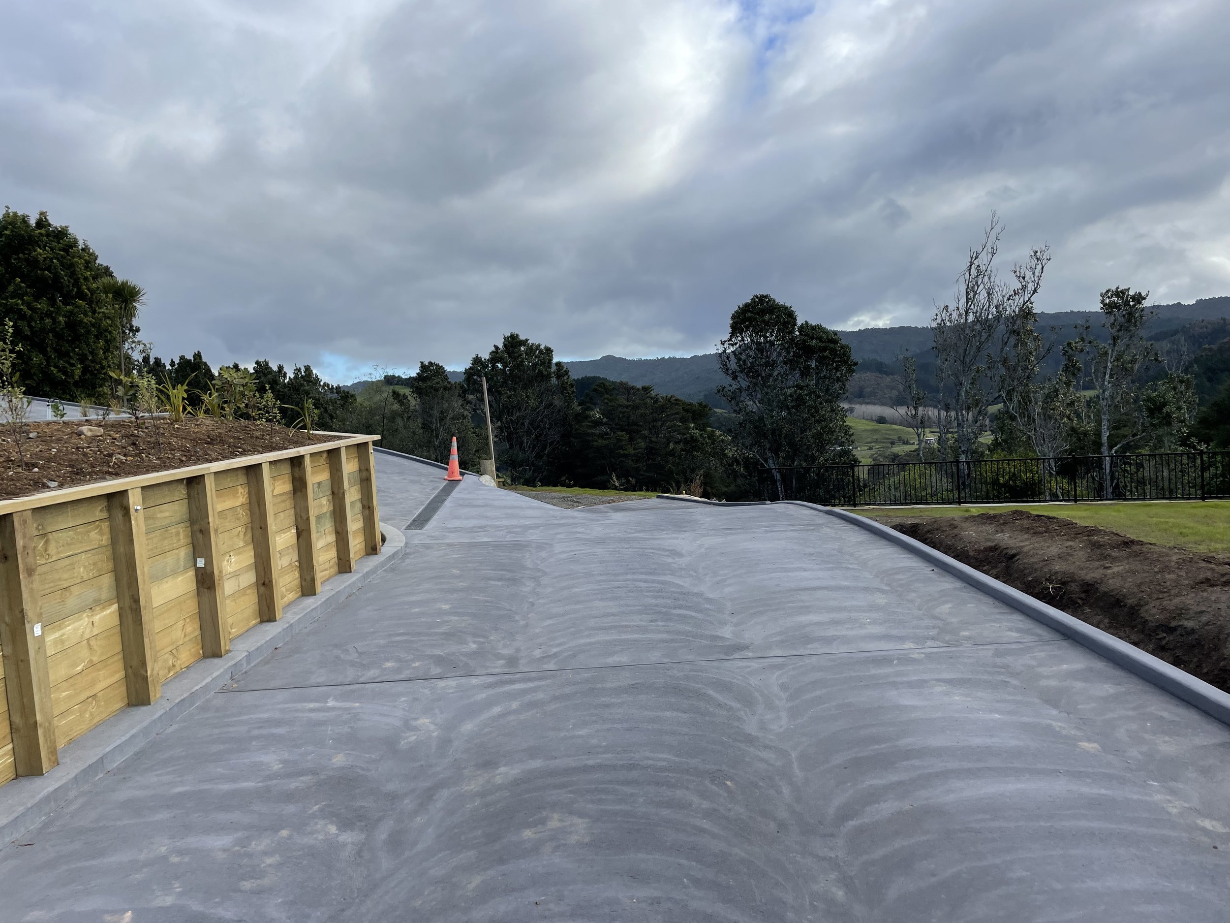 Newly paved, curved concrete pathway in a scenic outdoor area with a wooden retaining wall on the left and trees in the background under a cloudy sky.