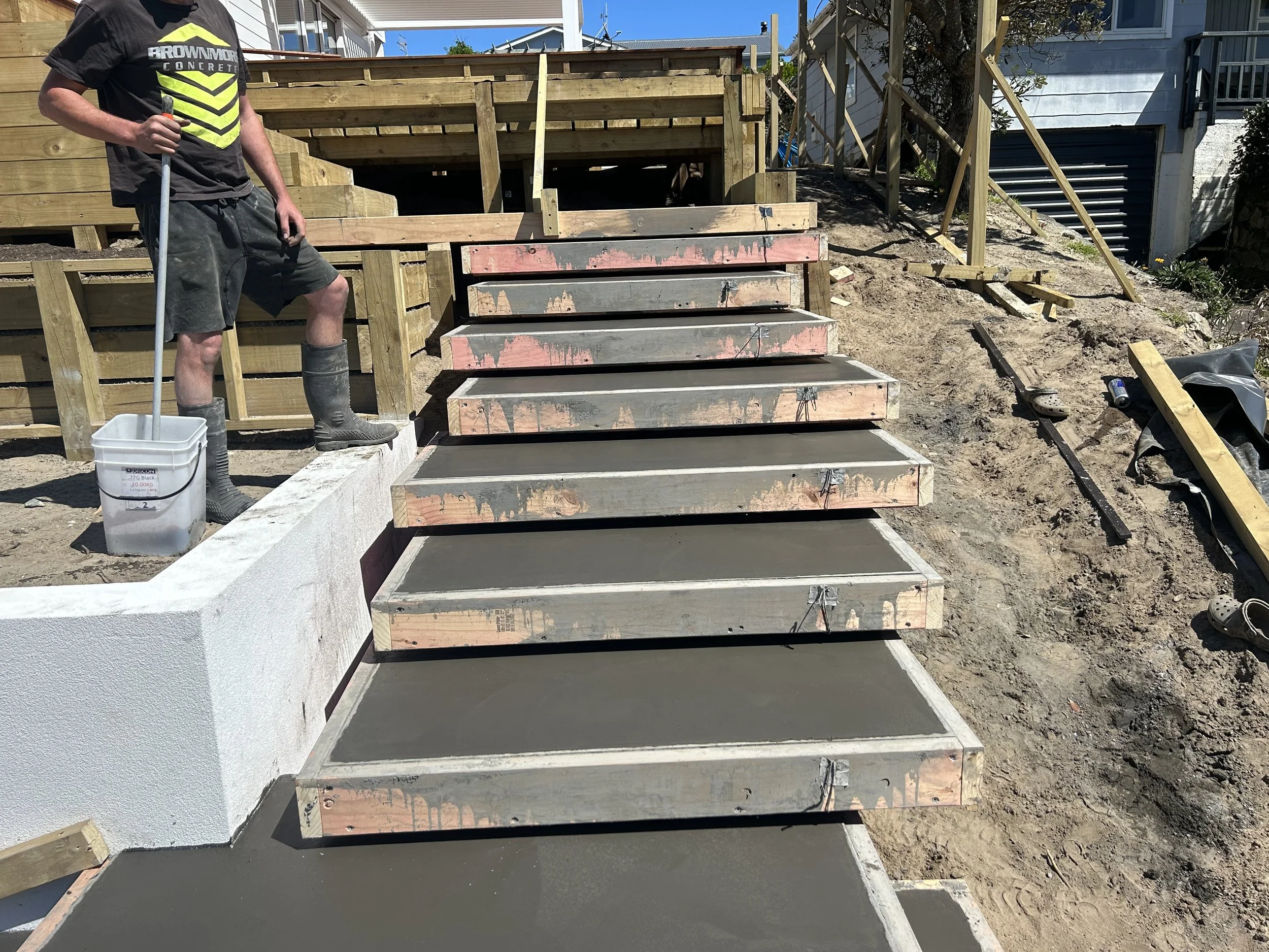 Construction worker standing next to a staircase under construction, with freshly poured concrete steps and wooden formwork in a backyard.