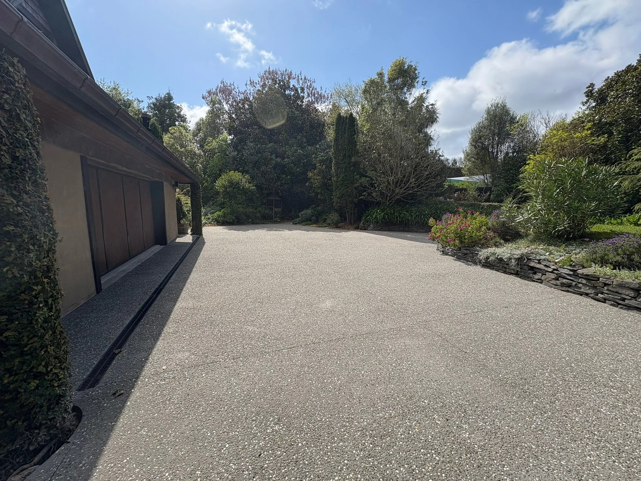 A spacious driveway with a gravel surface, bordered by lush green bushes and trees, with a portion of a house visible on the left and a cloudy blue sky overhead.