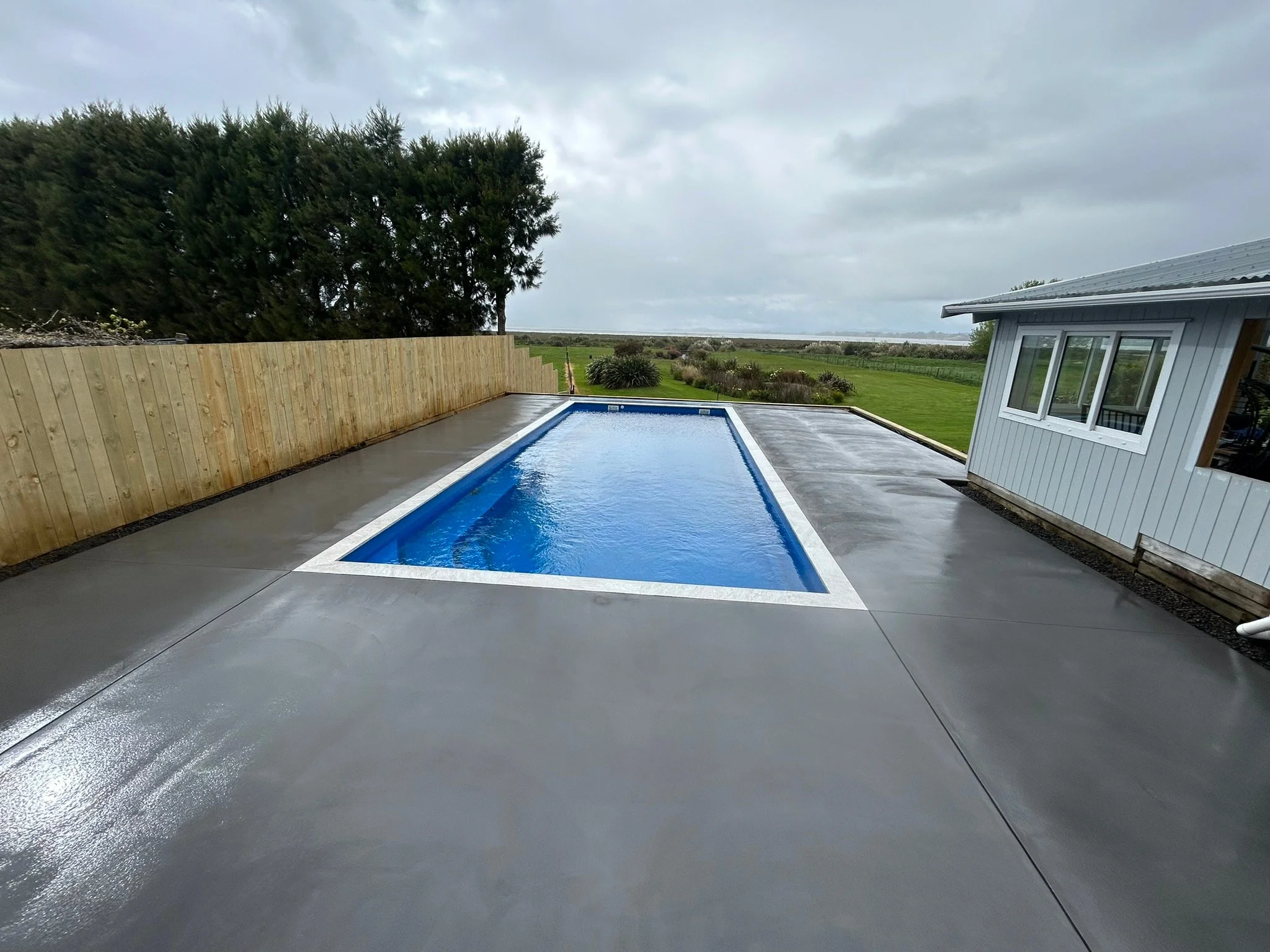 A rectangular swimming pool with blue water, surrounded by a concrete patio, next to a white house with a large window, and a wooden fence on the left side. Trees and open land are in the background, under a cloudy sky.