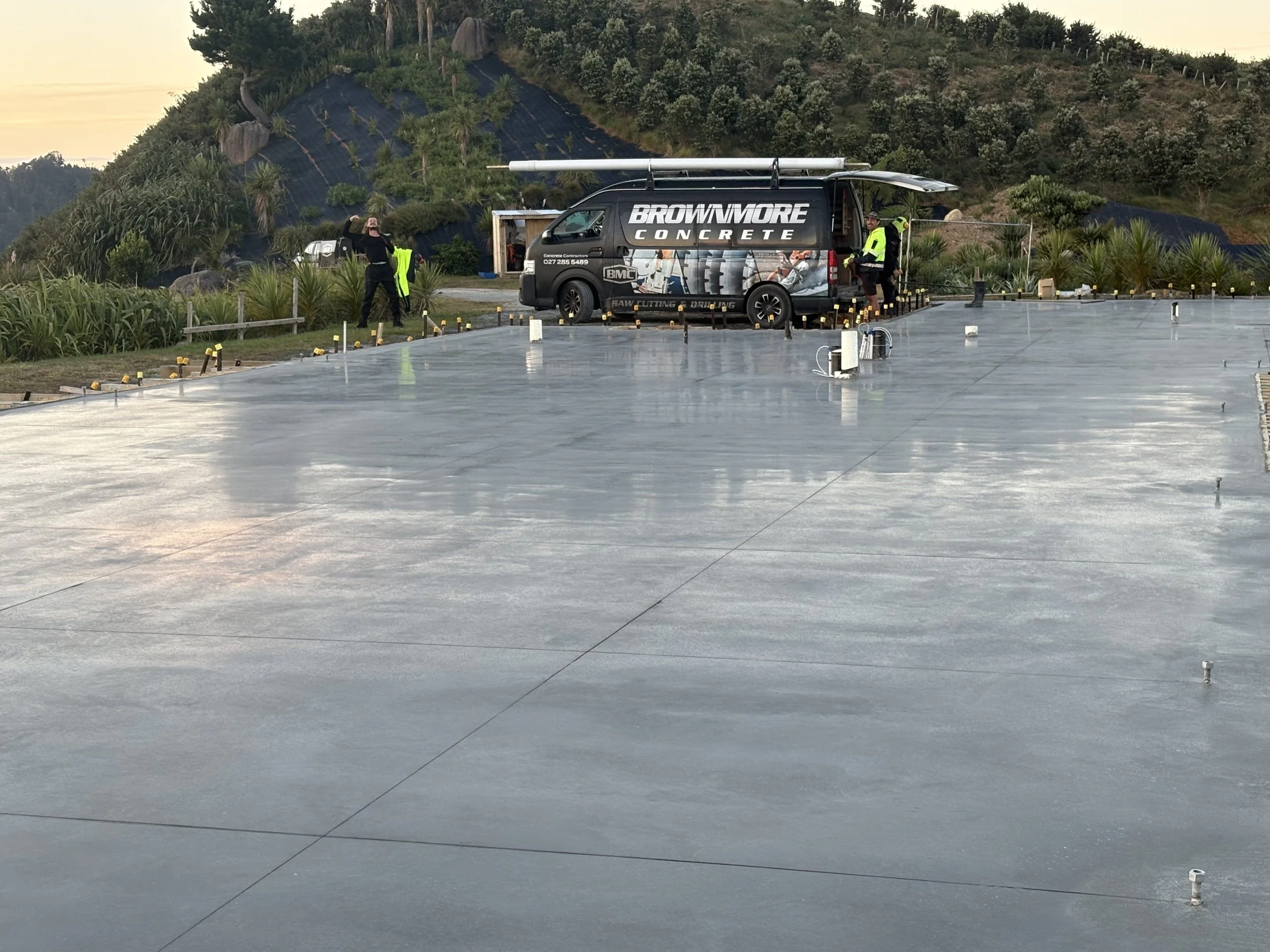 Construction workers are installing a large, smooth concrete slab on a building site with a scenic, hilly background and a decorative landscape. A black van with 'BROWIMORE CONCRETE' written on it is parked nearby.