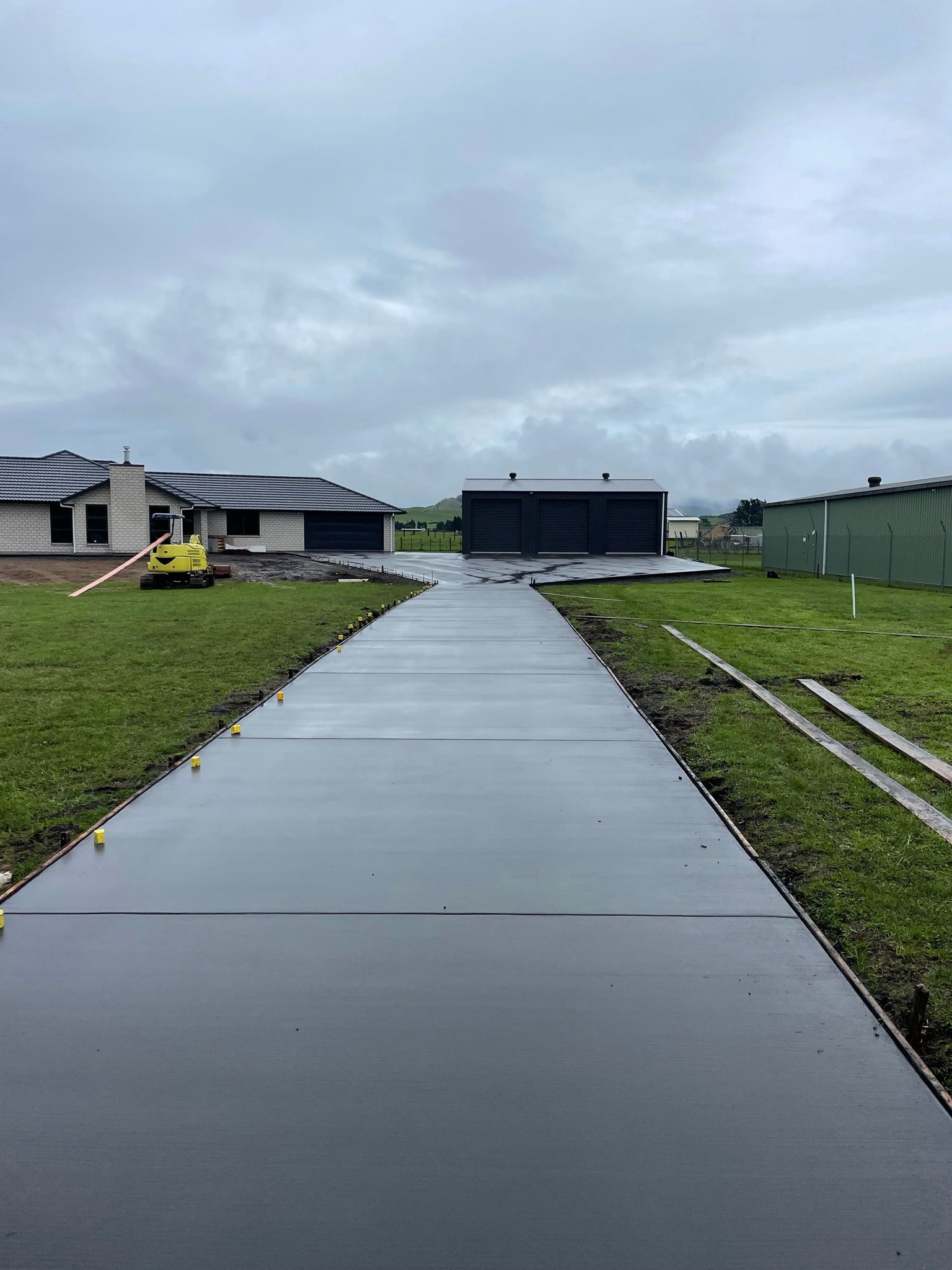 Newly poured concrete driveway in front of a house under construction, with construction equipment and a grassy yard around.