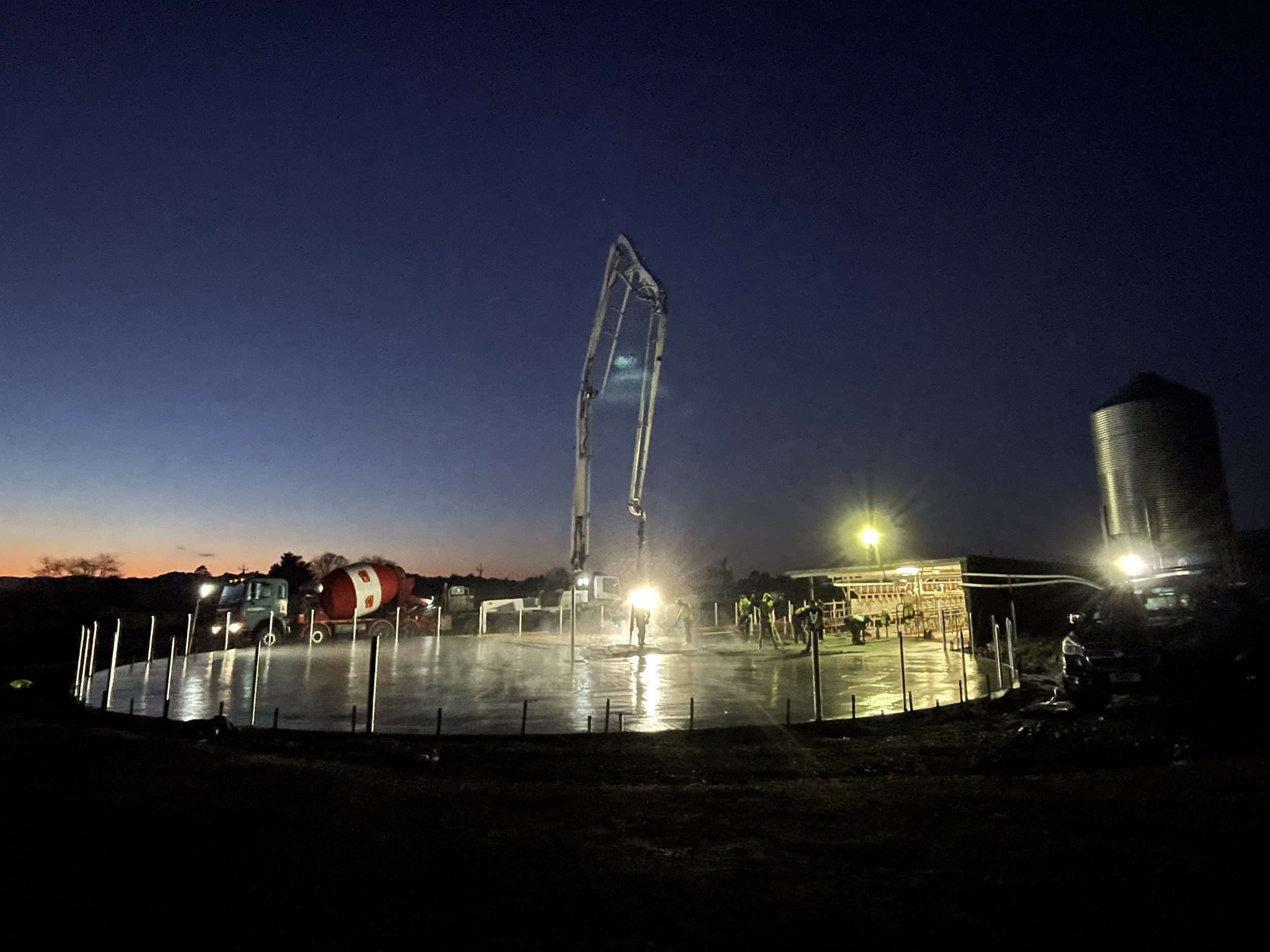Concrete slab being poured at a construction site during dusk or dawn with construction workers, a truck with a mixer, and a crane.