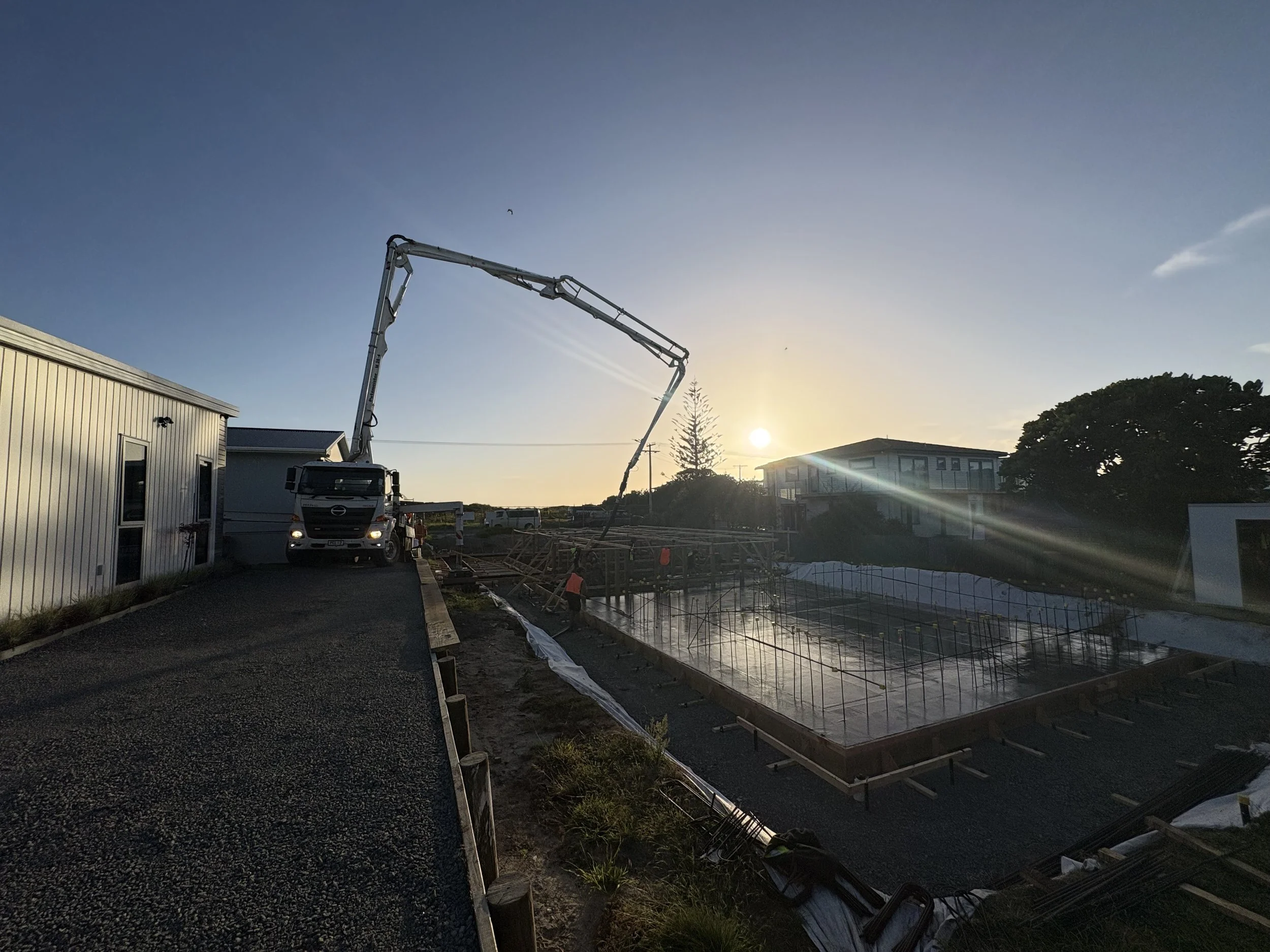 Construction site at sunset with a concrete pump truck pouring concrete for a building foundation, surrounded by construction workers and materials.