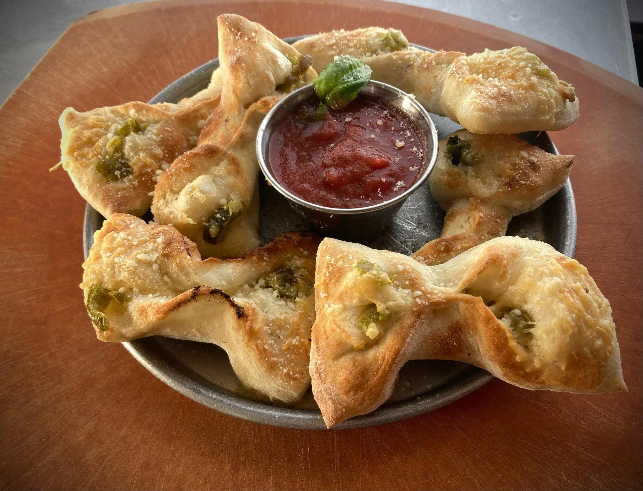 Plate of seven filled samosas with jalapeño bits, served with a small cup of red dipping sauce and a green chili on a round wooden plate.