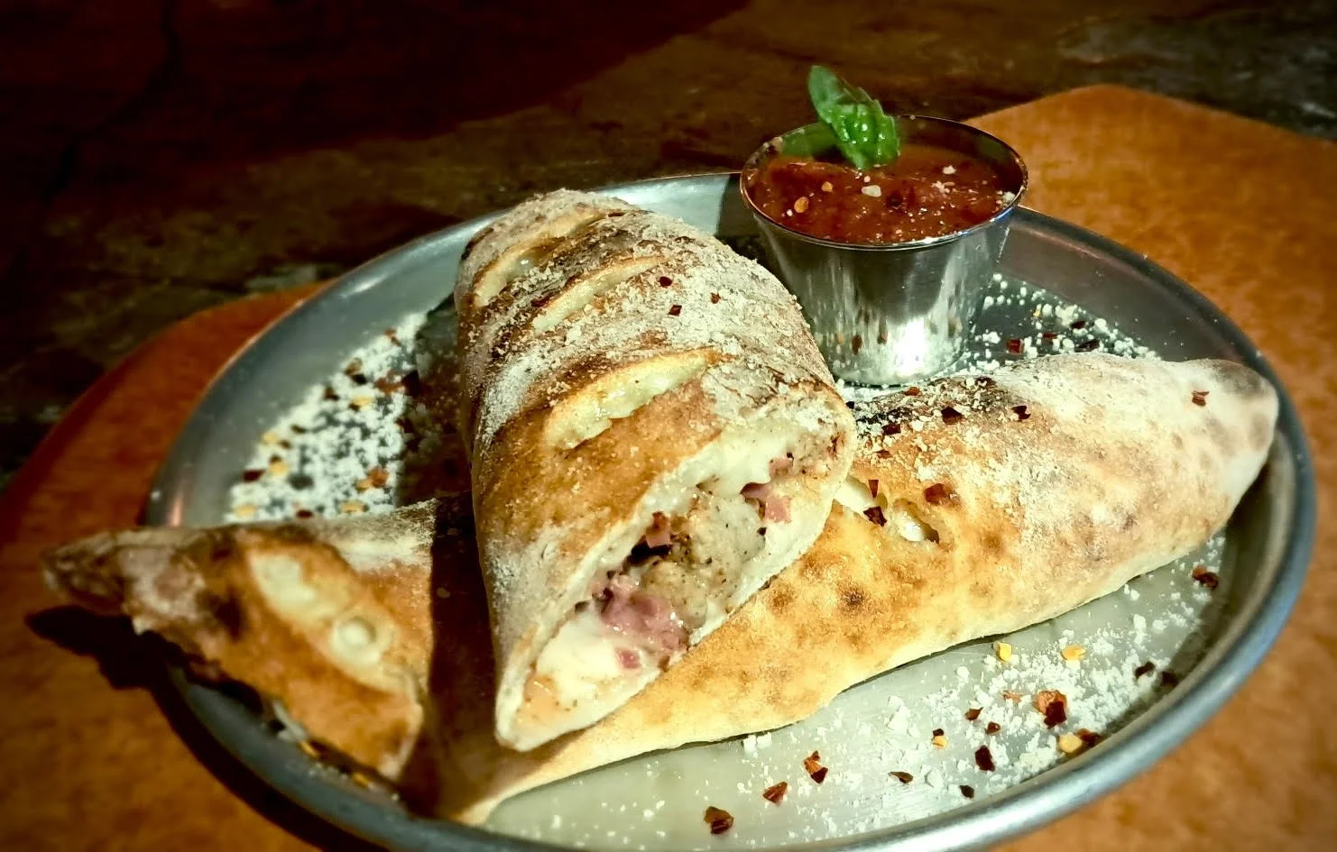 Plate with rolled savory pastry containing ham and cheese, sprinkled with powdered sugar, served with a side of red salsa in a small metal cup, garnished with a green leaf.