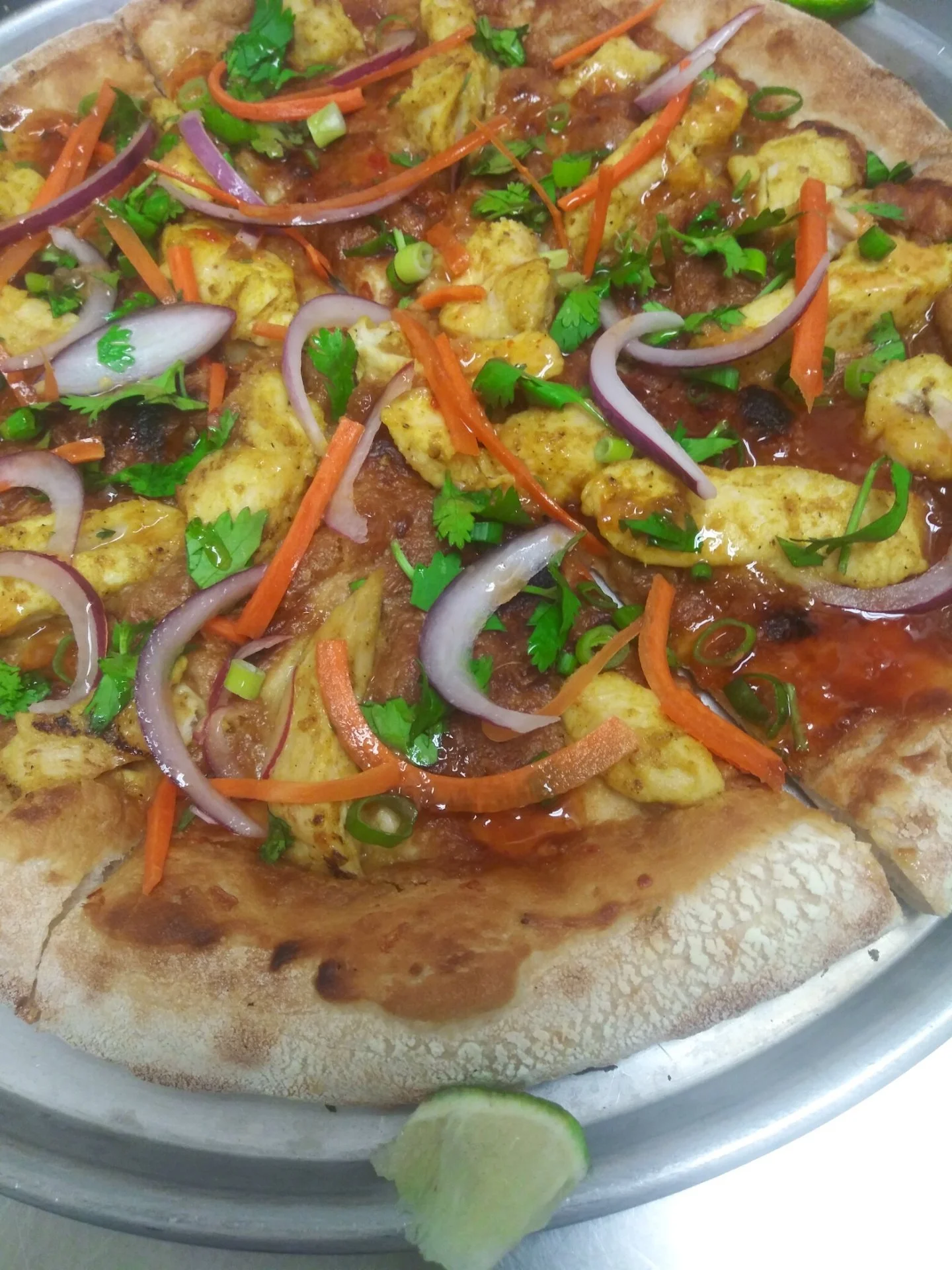 A pizza topped with chicken pieces, sliced red onions, shredded carrots, chopped green onions, cilantro, and a tomato-based sauce on a metal tray with lime wedge.