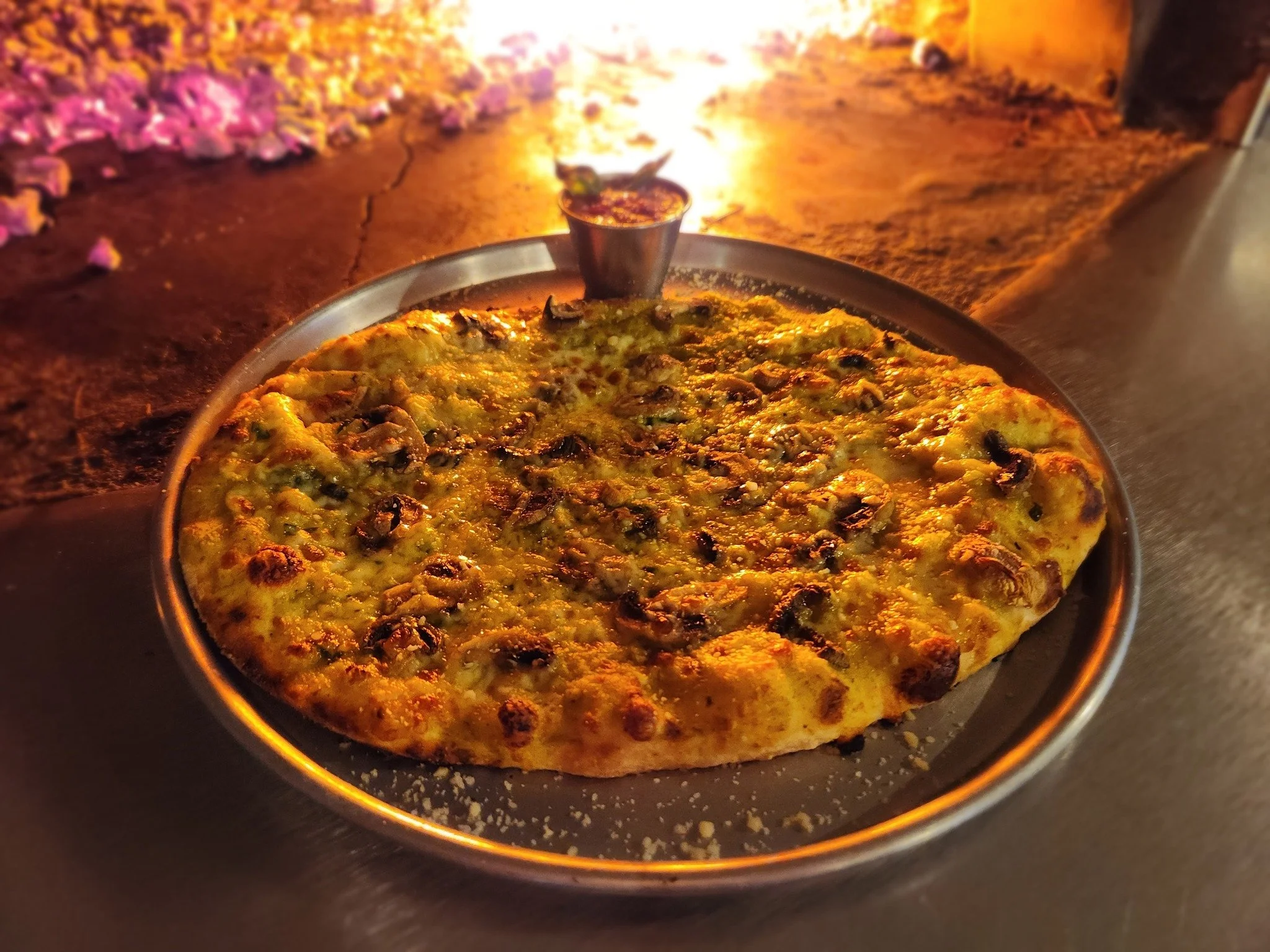 A pizza with cheese and pepperoni on a round metal tray, resting on a dark table, with a small cup of dipping sauce behind it, illuminated by warm lighting.