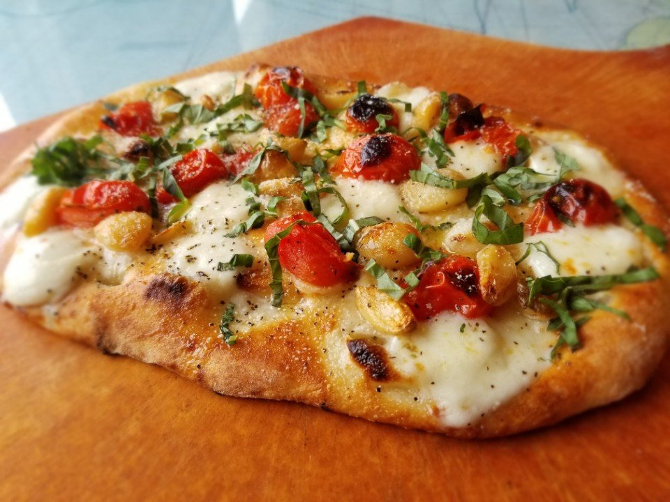 Close-up of a personal pizza on a wooden serving board, topped with tomatoes, basil, cheese, and grilled chicken pieces.