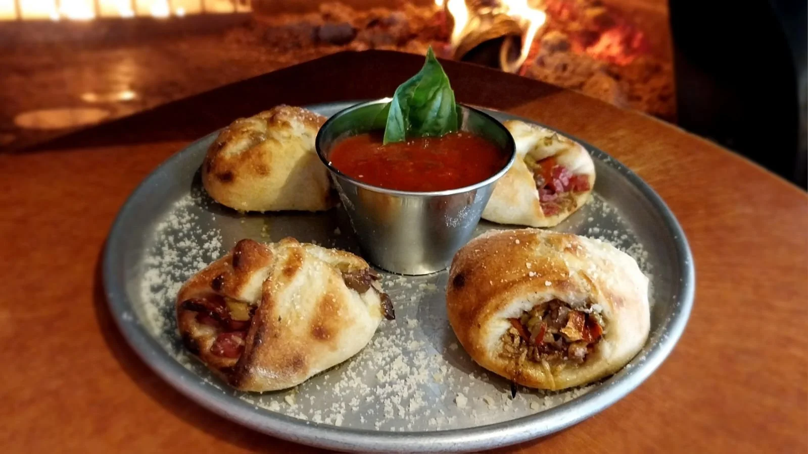 A round metal plate with four stuffed bread rolls, a small cup of marinara sauce topped with a basil leaf, and a dusting of grated cheese, with a fire in the background.