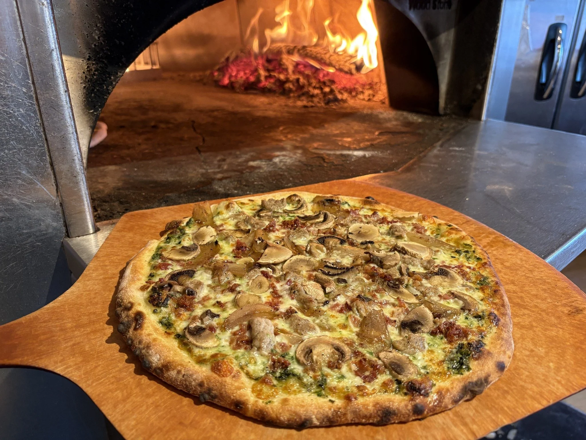 A freshly baked mushroom and cheese pizza on a wooden pizza peel being placed into a wood-fired pizza oven.