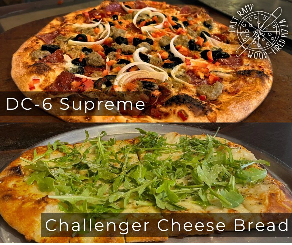 A pizza topped with onions, black olives, tomatoes, sausage, and pepperoni, served on a wooden board with a restaurant logo in the corner, next to a green salad pizza with arugula on top on a silver tray.