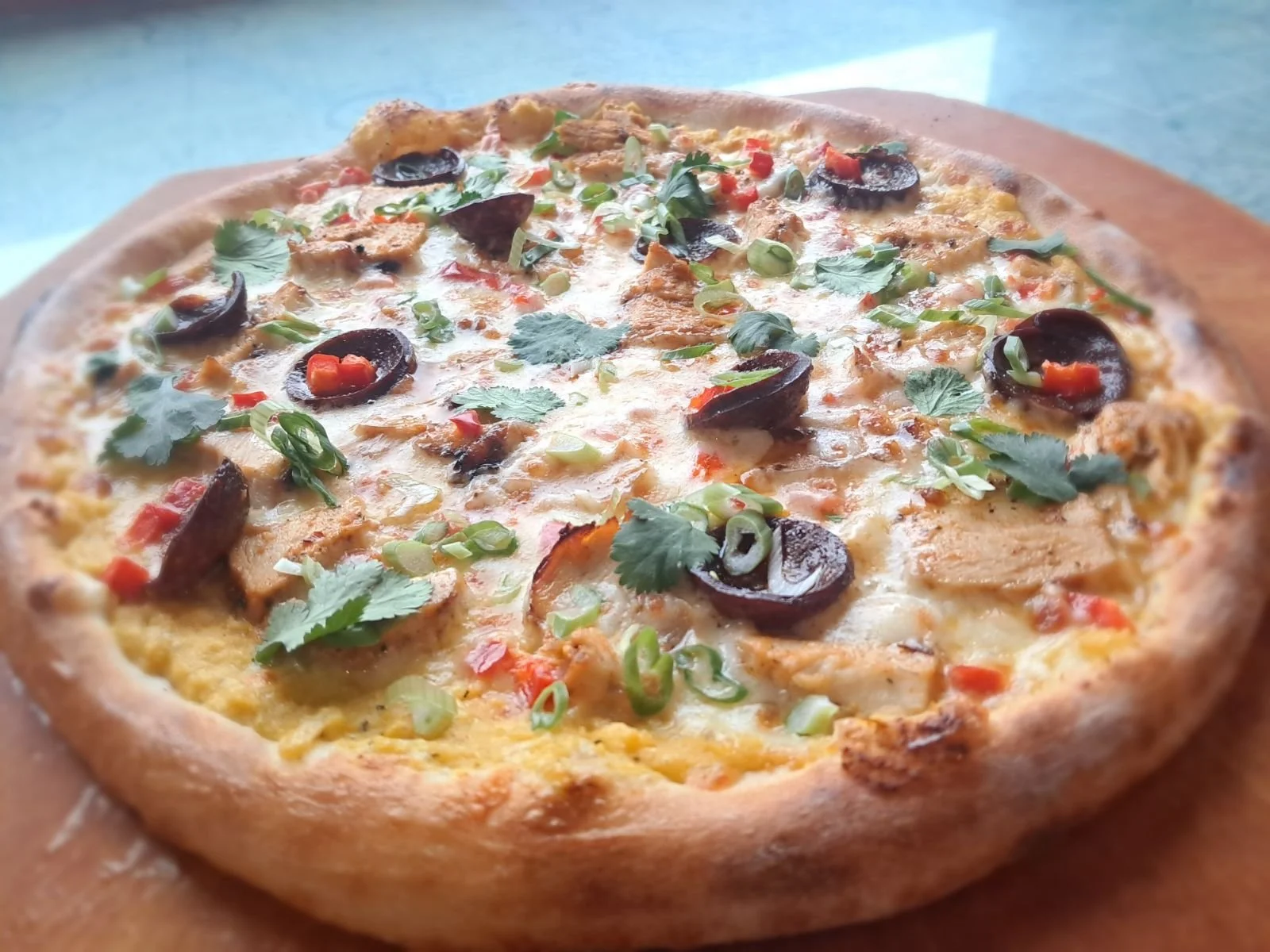 A close-up of a pizza with cheese, black olives, chopped green onions, cilantro, red bell peppers, and pieces of chicken on a wooden board.